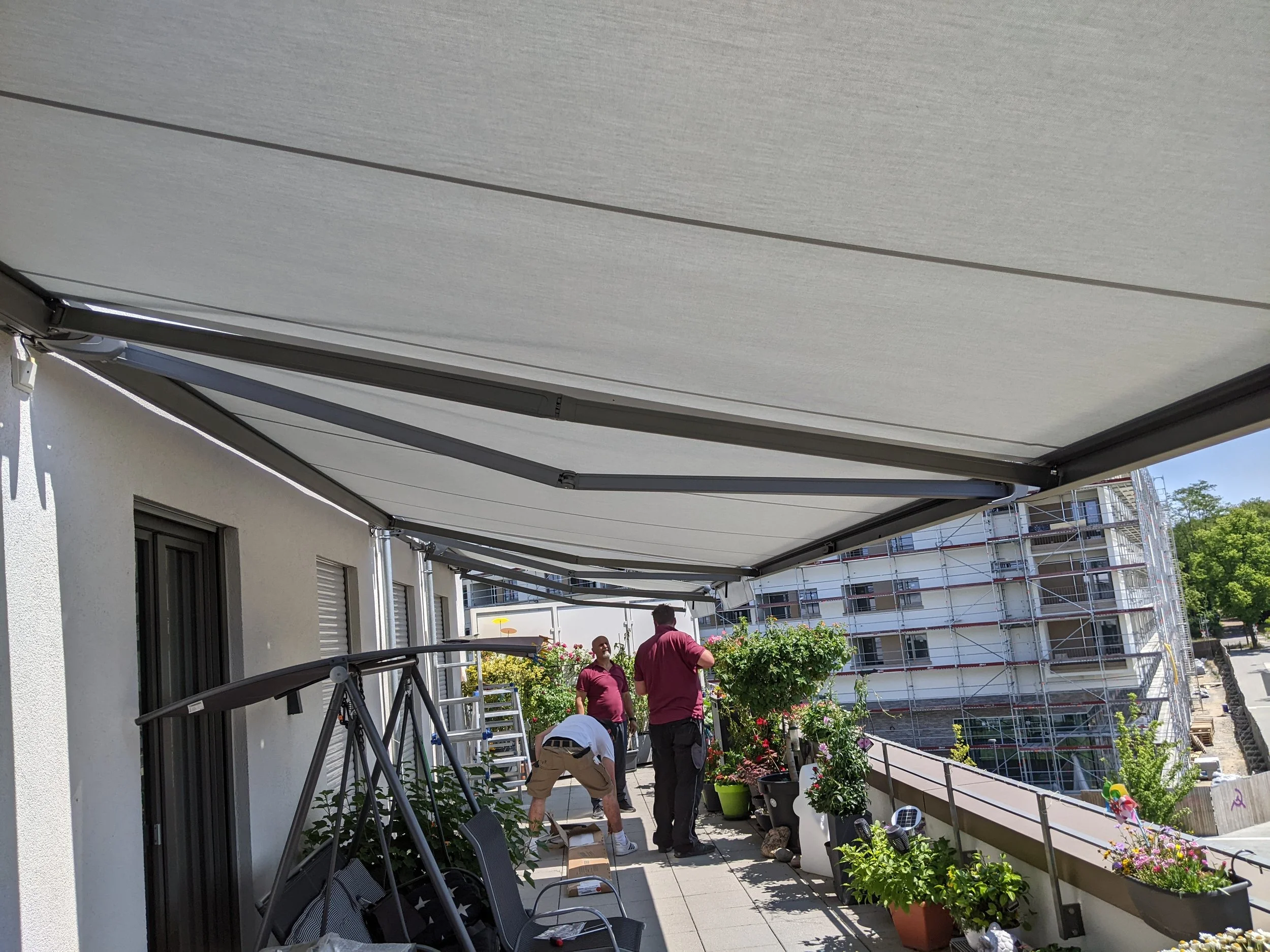 Menschen, die auf einer Balkon arbeiten, mit Pflanzen und Gartengeräten, im Hintergrund ein Gebäude, das mit Gerüsten gebaut wird.