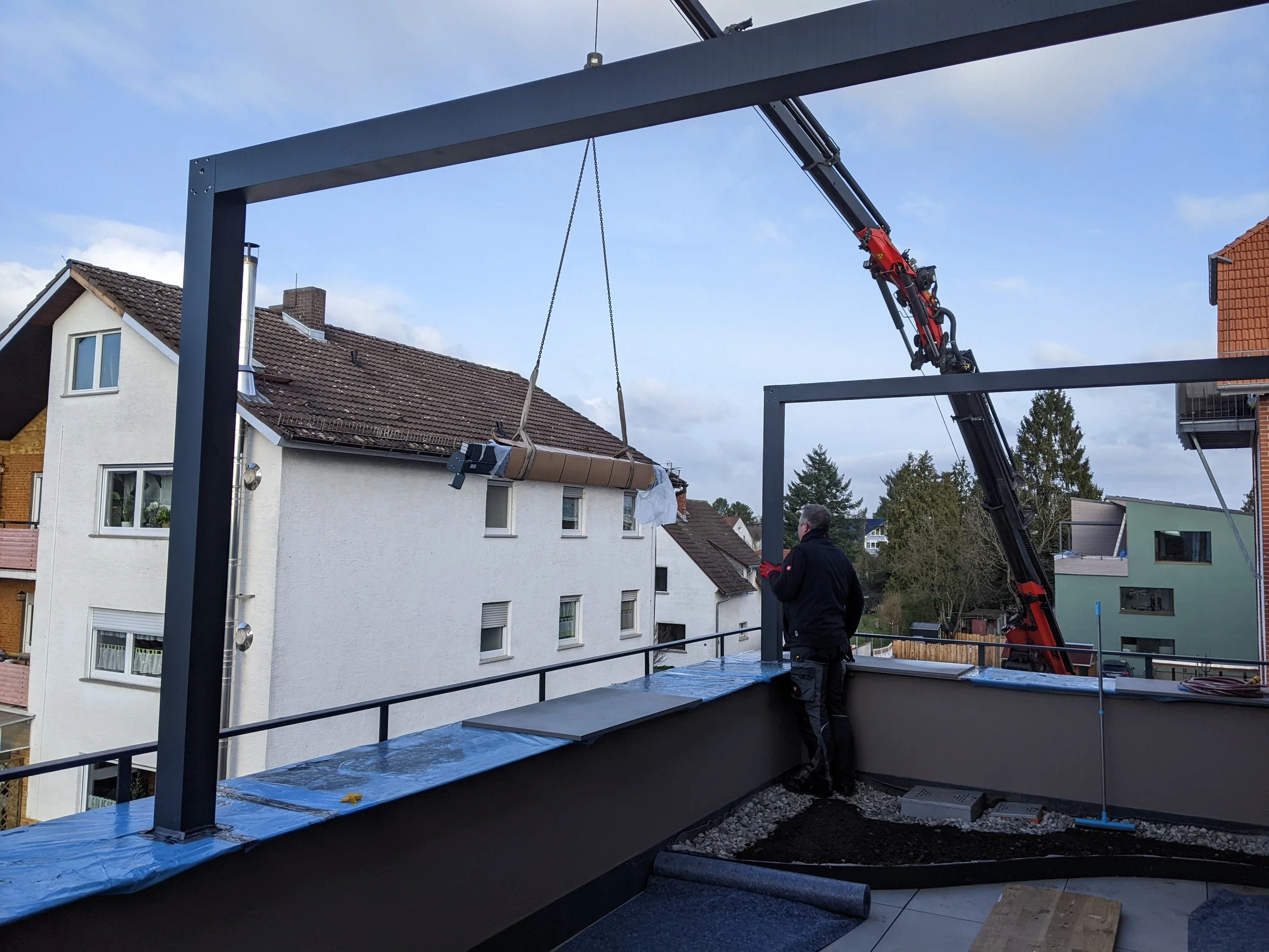 Ein Mann steht auf einem Balkon und beobachtet, wie mit einem Kran eine große Holzrolle auf den Balkon gehoben wird.