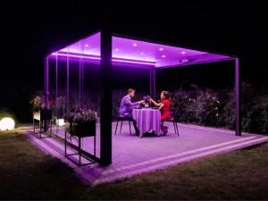 Zwei Menschen sitzen an einem Tisch unter einer beleuchteten Pergola im Freien bei Nacht.