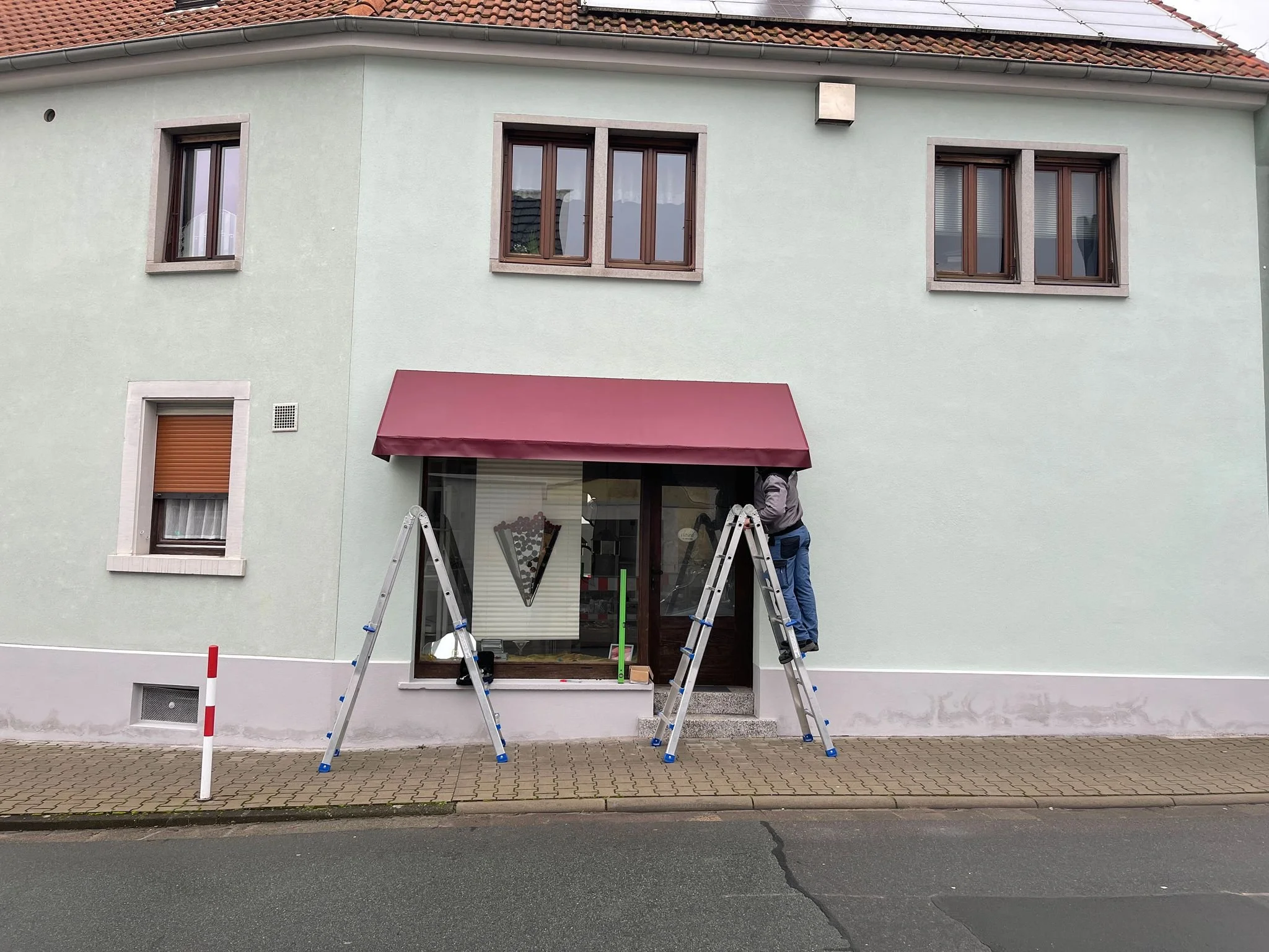 Mann arbeitet an einem Fenster eines Gebäudes mit einem roten Markise. Er steht auf einer Leiter und benutzt Werkzeuge.