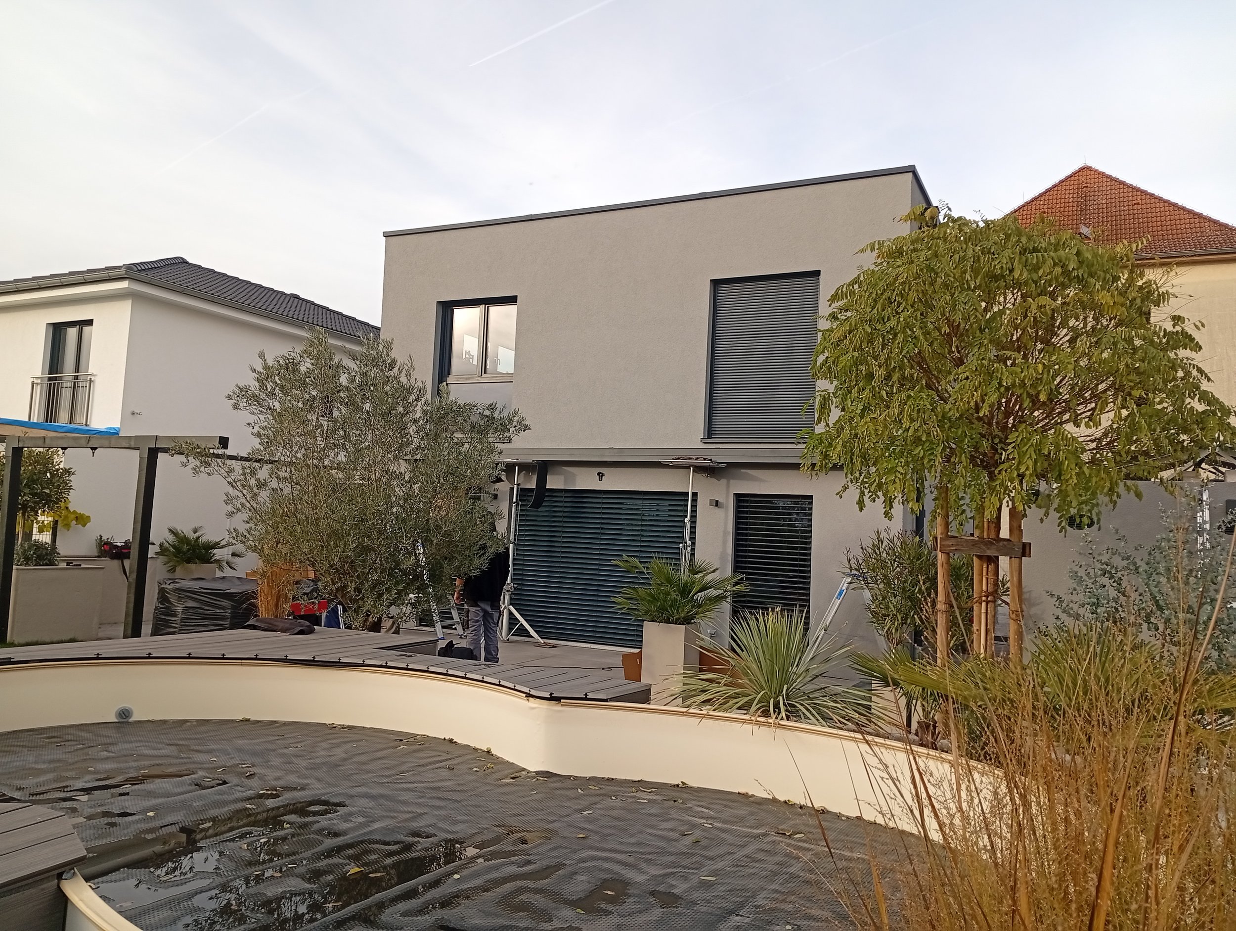 Moderne zweistöckige Villa mit Gartenterrasse, umgeben von Pflanzen und Bäumen, bei Sonnenuntergang.