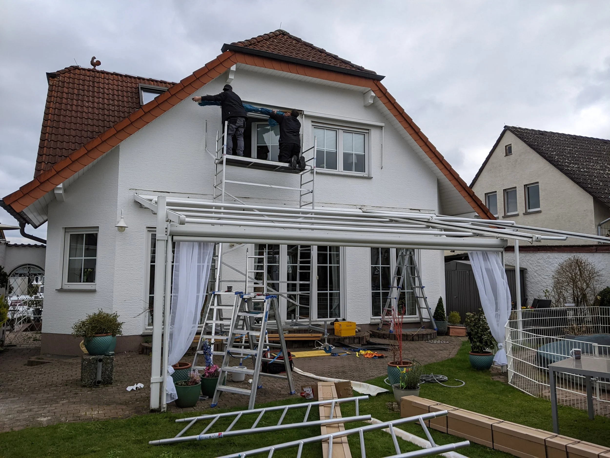 Zwei Männer arbeiten an einem Gerüst vor einem weißen Haus mit rotem Ziegeldach, dabei montieren sie eine Plane oder Abdeckung im oberen Bereich der Fassade.