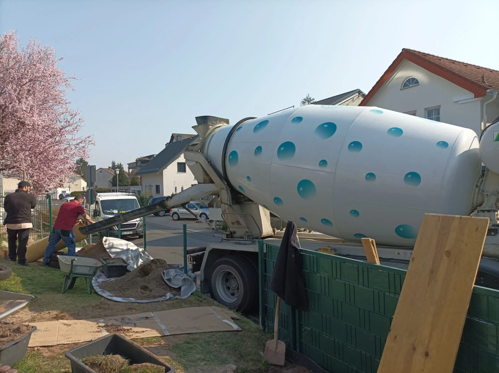Zementmischer auf einer Baustelle mit zwei Arbeitern, umgeben von Baumaschinen und Baumaterialien.