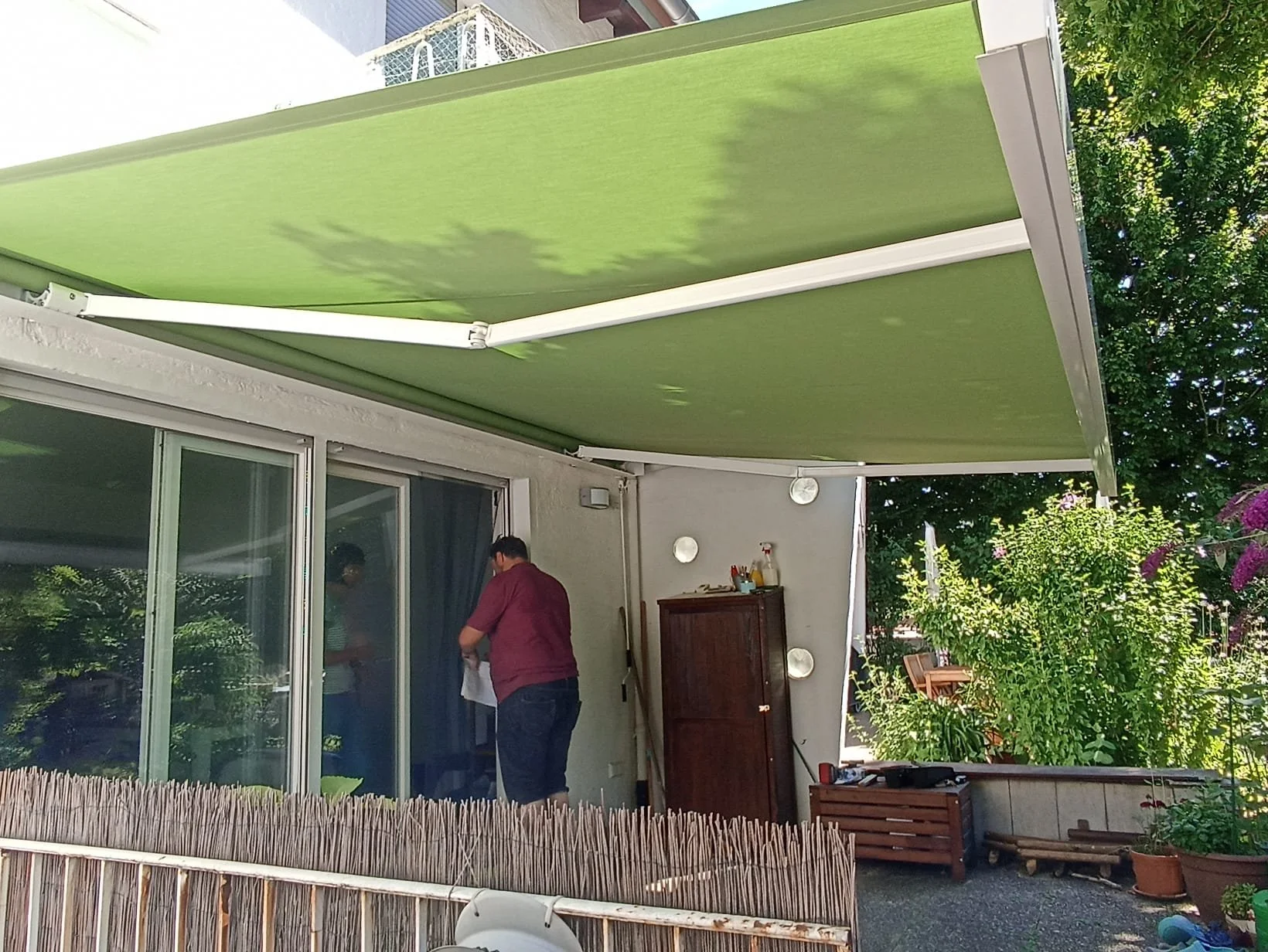 Ein Mann steht auf einer Terrasse vor einem Glastür, die zu einem Haus führt, mit einer grünen Markise darüber. Es gibt Pflanzen und Gartenmöbel im Hintergrund.