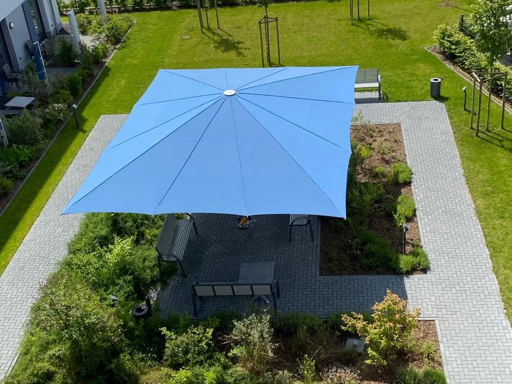 Vogelperspektive auf einen grünen Garten mit einem Blütenteich, einem Pavillon mit blauer Sonnenschirm, sowie Gartenmöbeln und Büschen.
