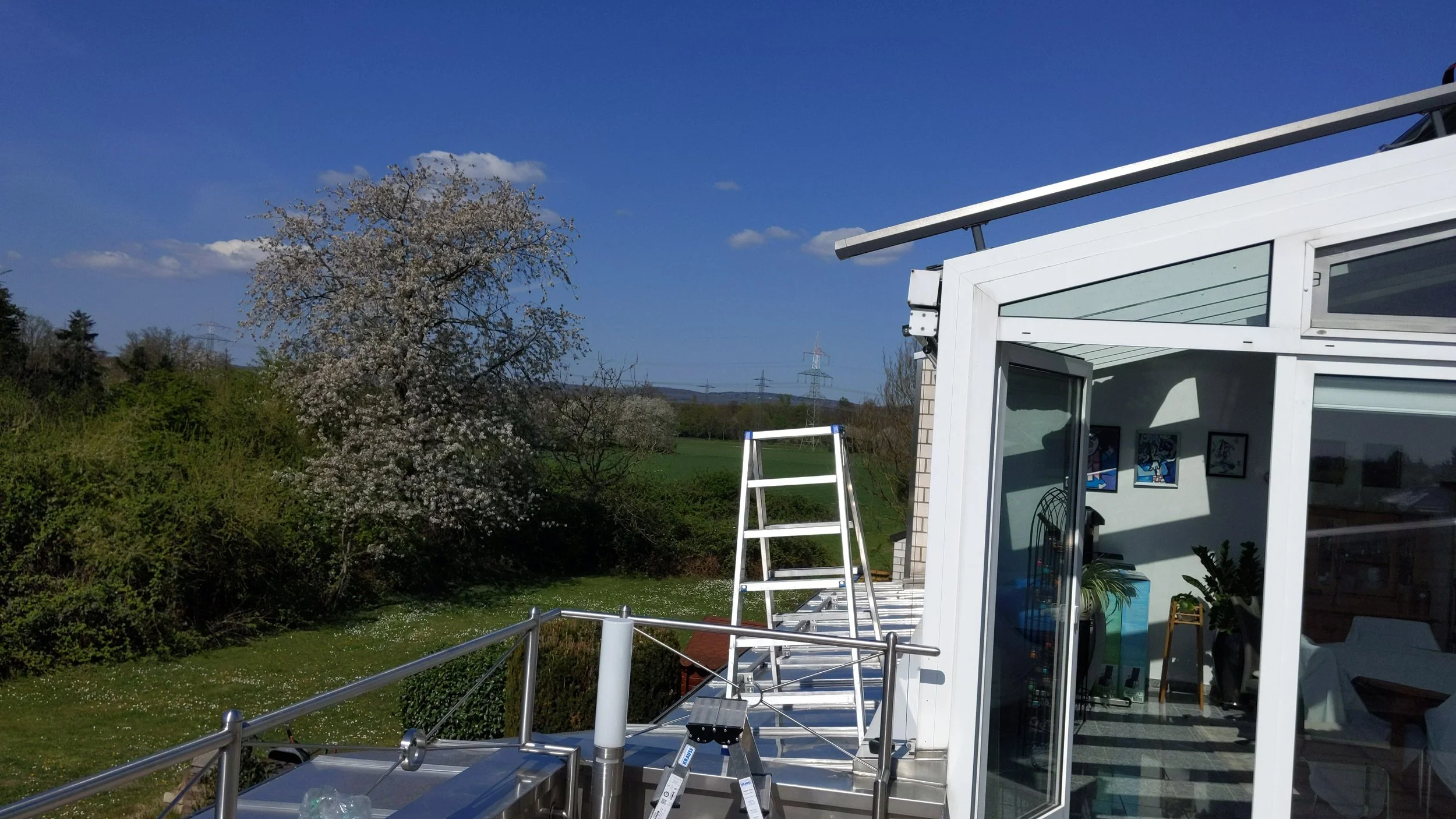 Blick auf eine Terrasse mit Leiter, umgeben von einem Haus mit großen Fenstern, im Hintergrund Bäume und ein blauer Himmel.