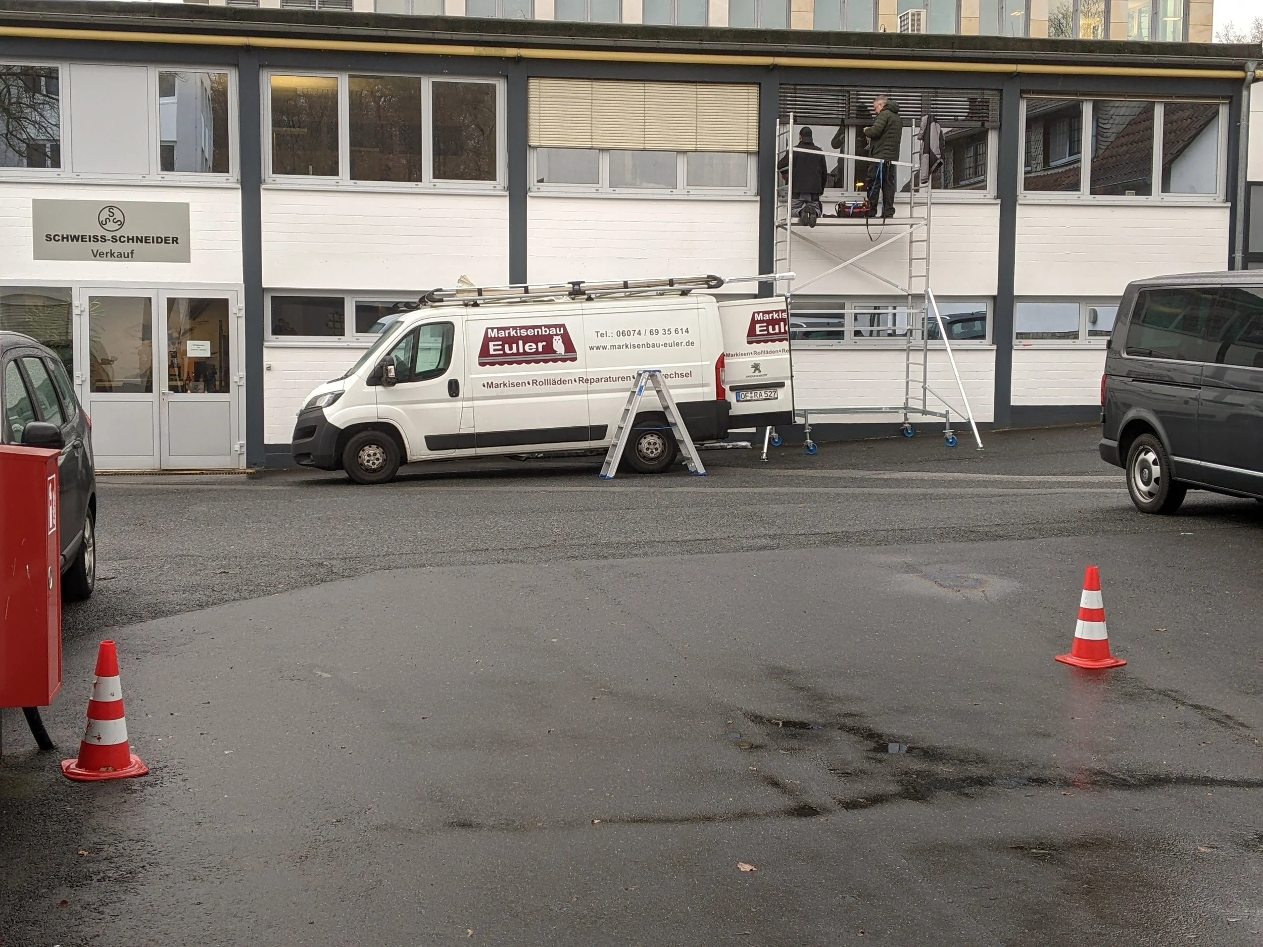 Arbeiten an der Außenfassade eines Gebäudes, mit Arbeitern auf einer Gerüsttreppe und einem weißen Firmenwagen vor dem Gebäude, das den Namen 'Markisenbau Euler' trägt.