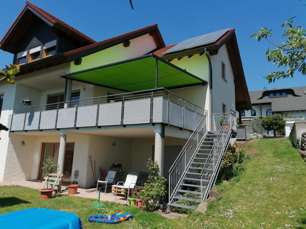 Ein mehrstöckiges Haus mit Balkon und Sonnenkollektoren auf dem Dach, im Garten sind Liegestühle, ein kleiner Pool und Spielzeug.