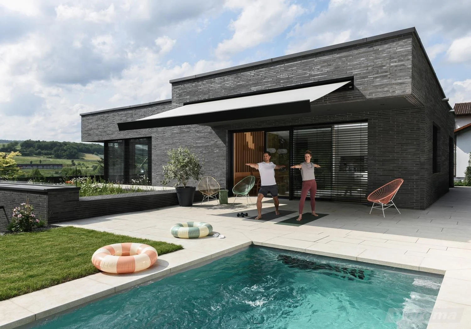 Zwei Menschen machen Yoga am Pool vor einem modernen schwarzen Haus mit großen Glasfenstern und Gartenmöbeln, in einer ländlichen Umgebung.