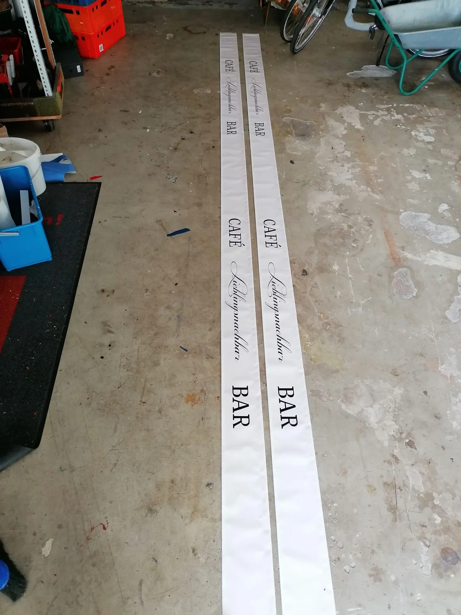 Zwei lange weiße Banner mit schwarzen und grauen Schriftzügen, die die Wörter 'CAFÉ', 'Bar', 'Kaffe' und 'Kaffeeautomaten' enthalten, liegen auf einem Boden in einer Garage oder Werkstatt.