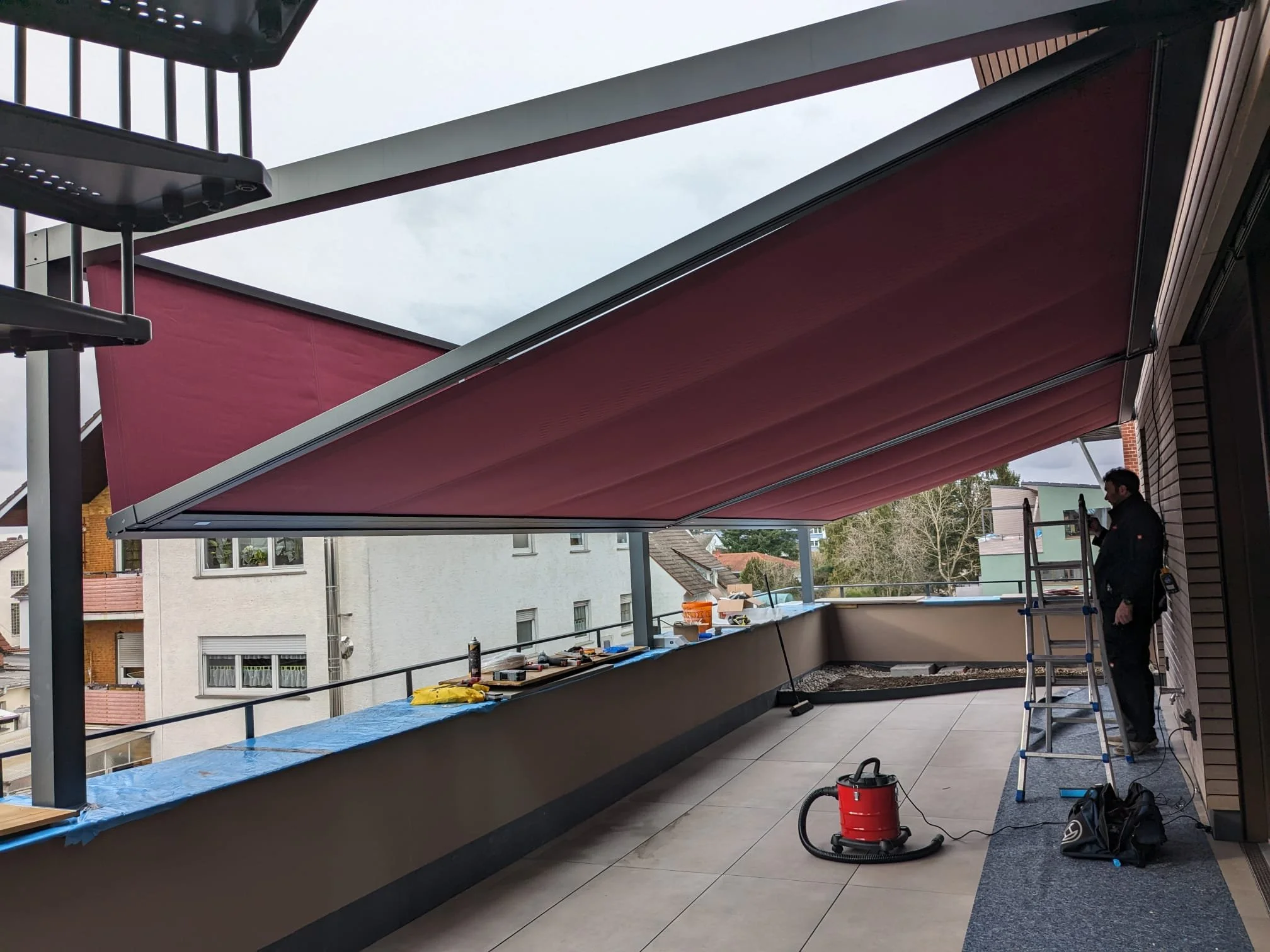 Ein Balkon mit einem roten Sonnenschutz, auf dem ein Mann mit Leiter und Werkzeug steht, während Arbeiten am Balkon durchgeführt werden.