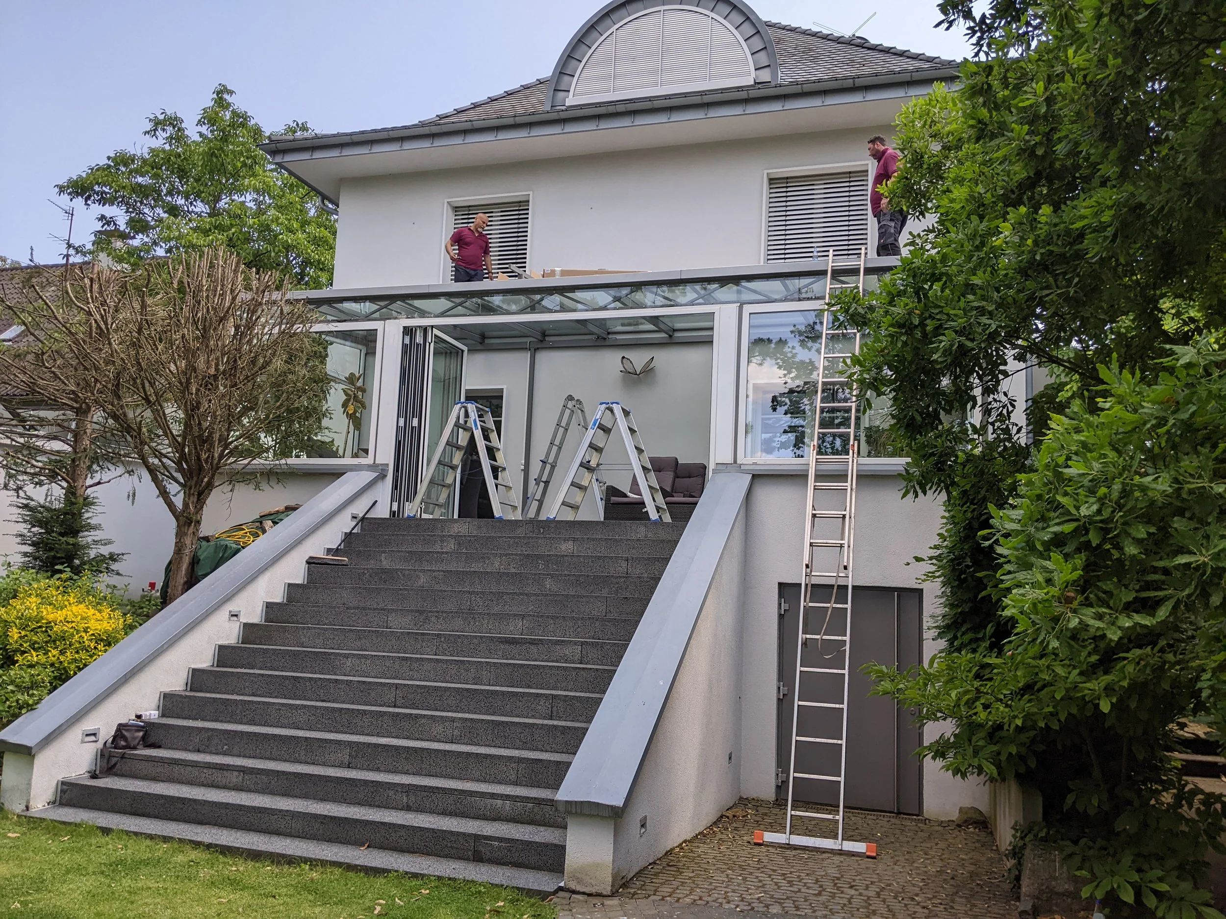 Zwei Männer stehen auf einem Balkon im Obergeschoss eines weißen Hauses, während auf der Terrasse im Erdgeschoss mehrere Alu-Leitern stehen. Es sind Leitern an der Hauswand positioniert, mit einer an die Wand gelehnten Leiter. Im Hintergrund sind Bäu
