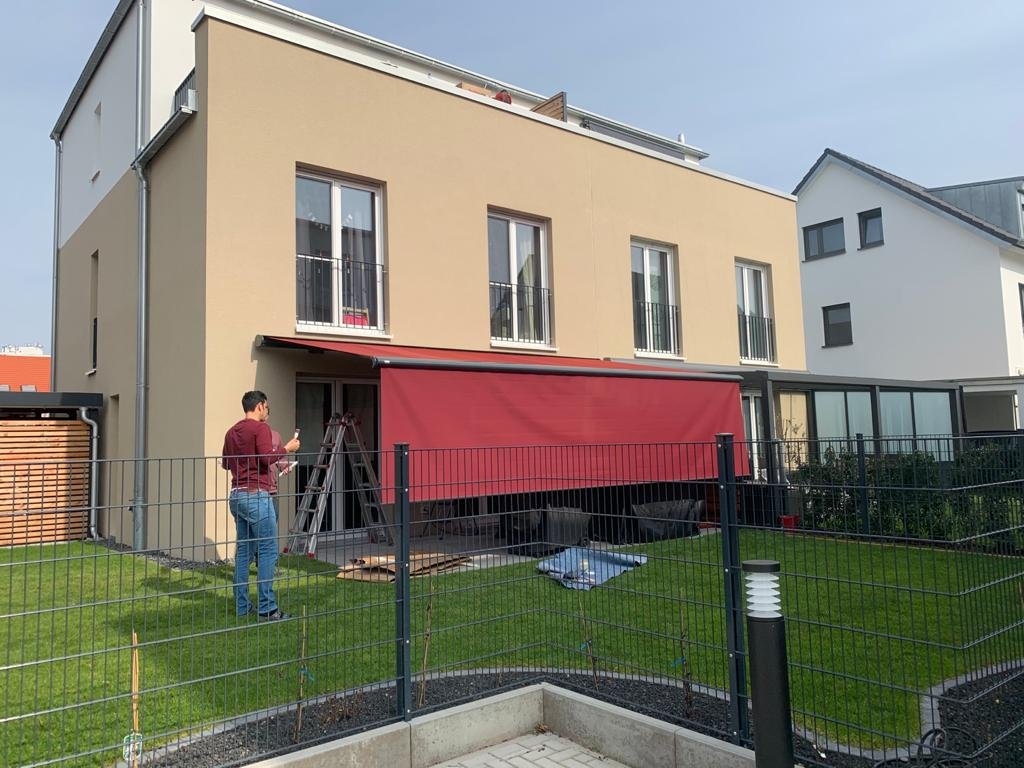Ein modernisiertes Mehrfamilienhaus mit einem Garten, in dem am Tag gearbeitet wird. Ein Mann steht vor einem Fenster mit einem roten Sonnenschutz und hält ein Smartphone in der Hand, während eine Leiter im Garten steht.