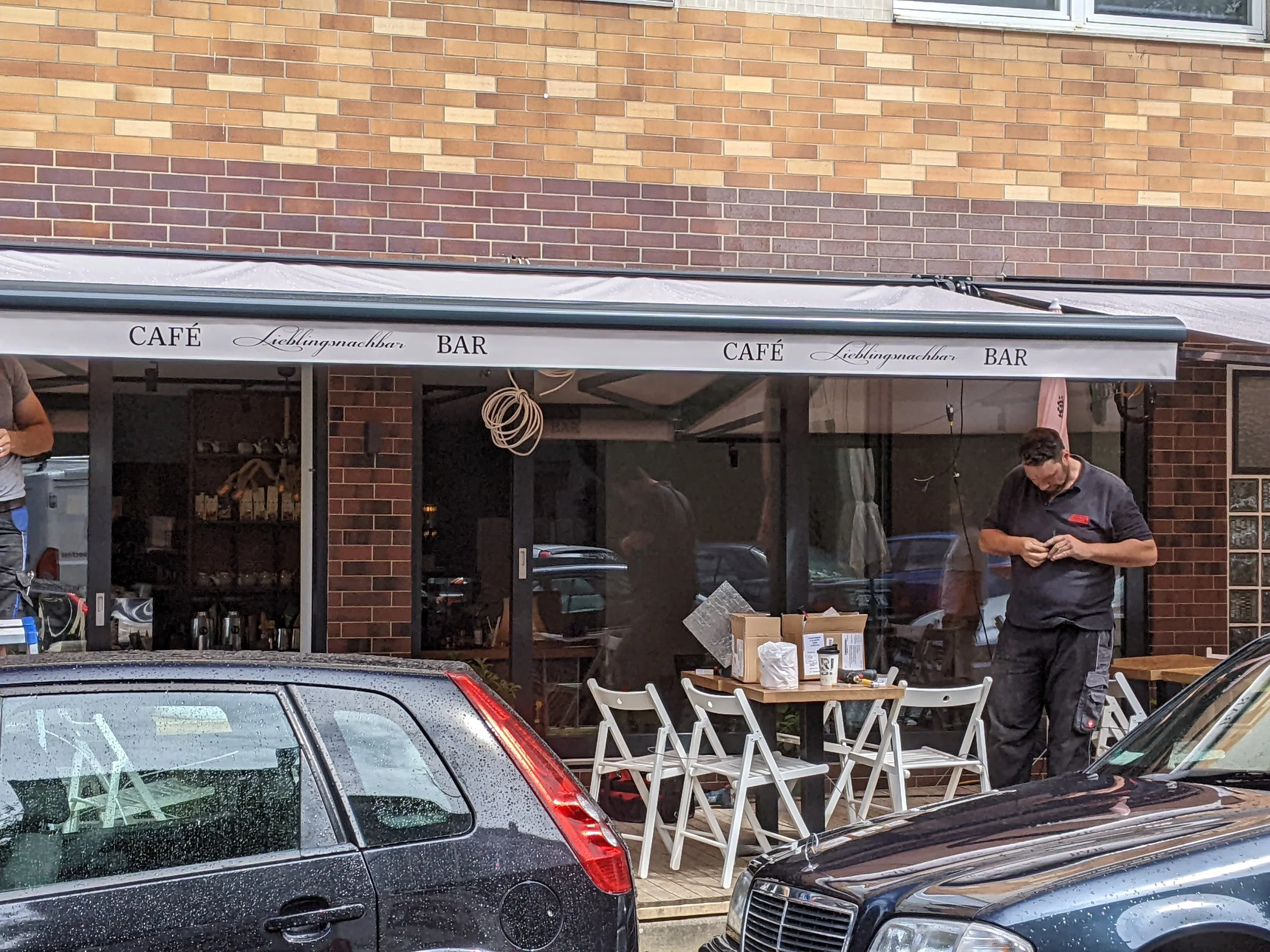 Außenansicht eines Cafés mit Sitzgelegenheiten, zwei Männer stehen draußen, regnerischer Tag, Autos vor dem Café.