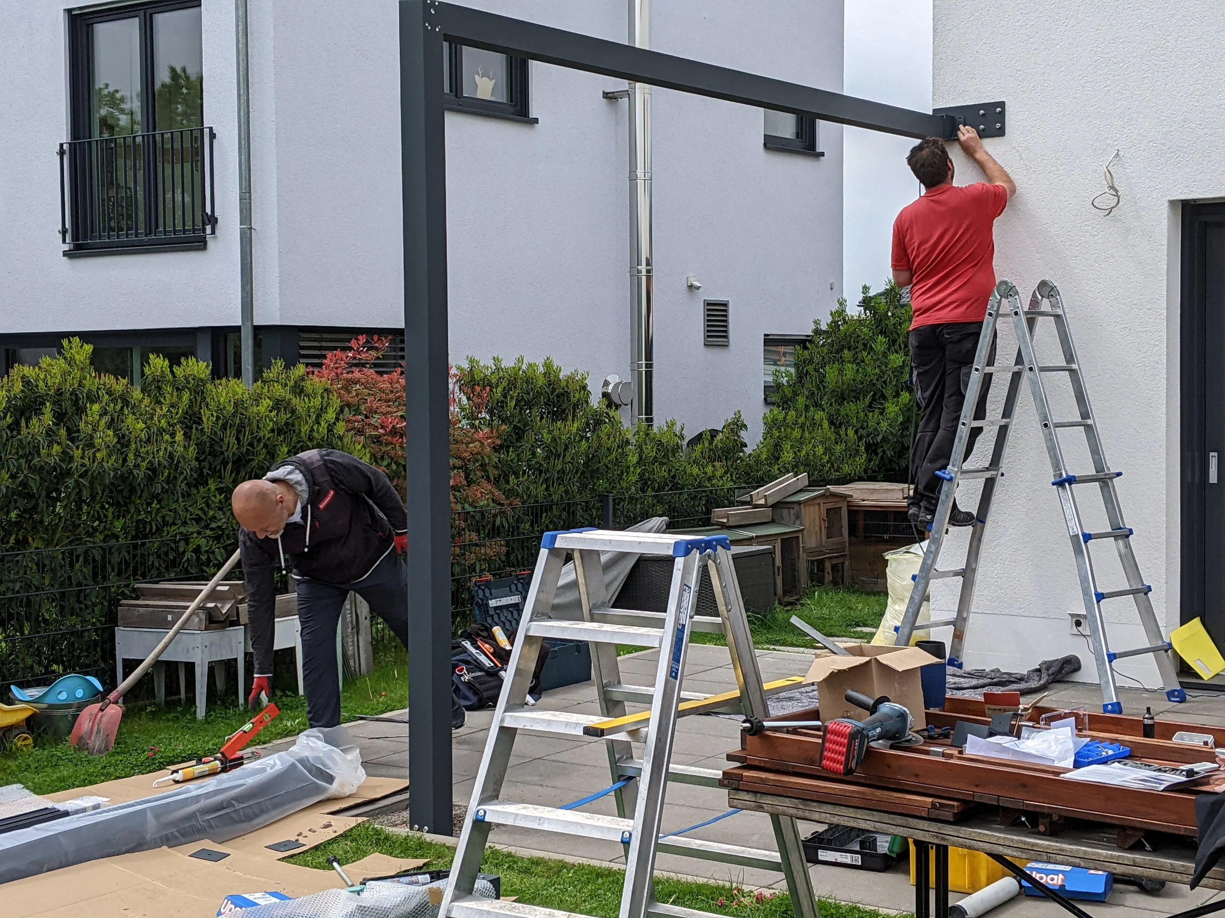 Zwei Männer bei der Installation eines Metallrahmens im Garten vor einem weißen Haus, umgeben von Werkzeugen und Baumaterialien.