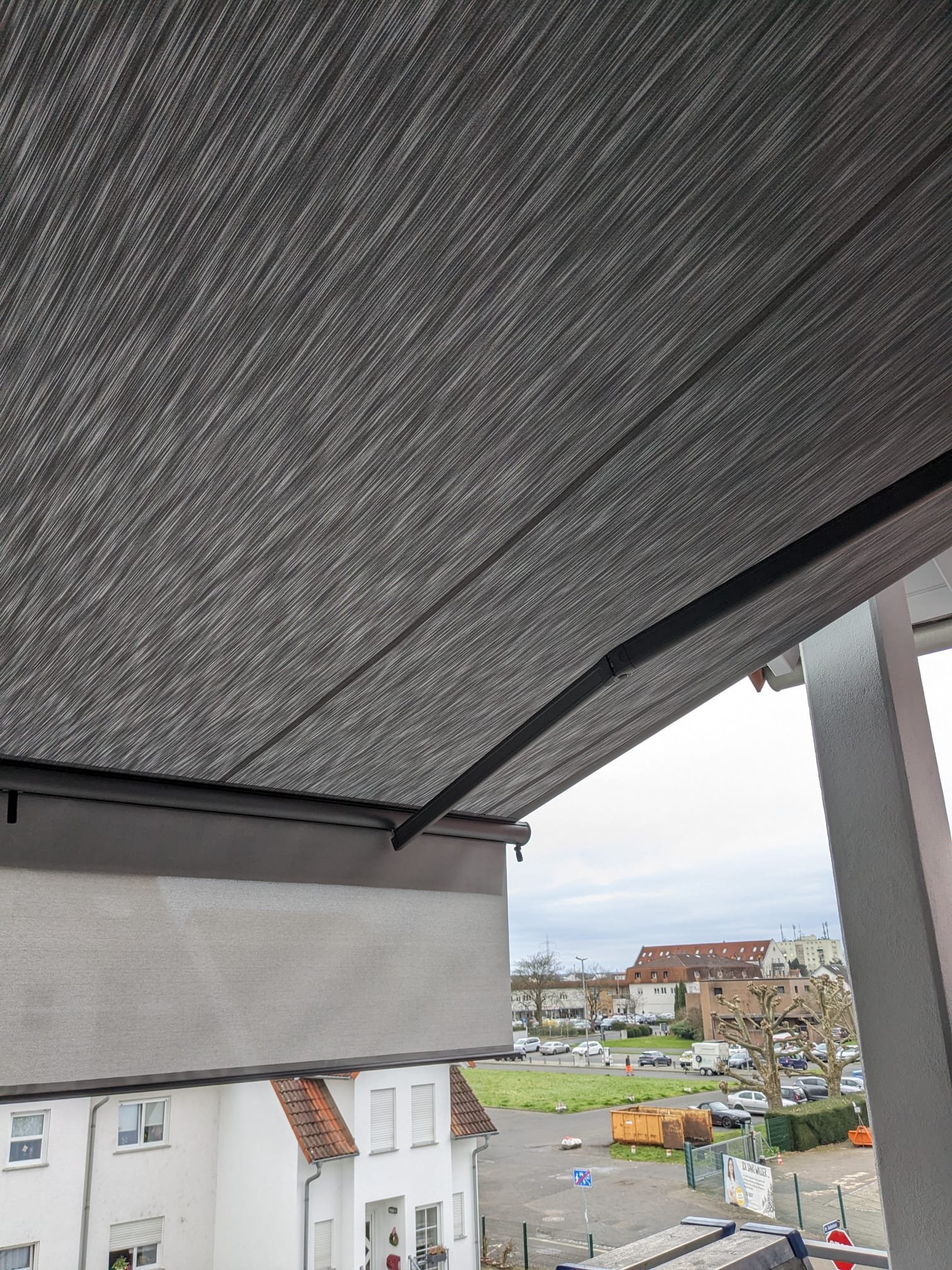 Balkon mit Holzdach, Blick auf eine Stadt mit weißen Häusern, Parkplätzen und Bäumen, bewölkter Himmel.