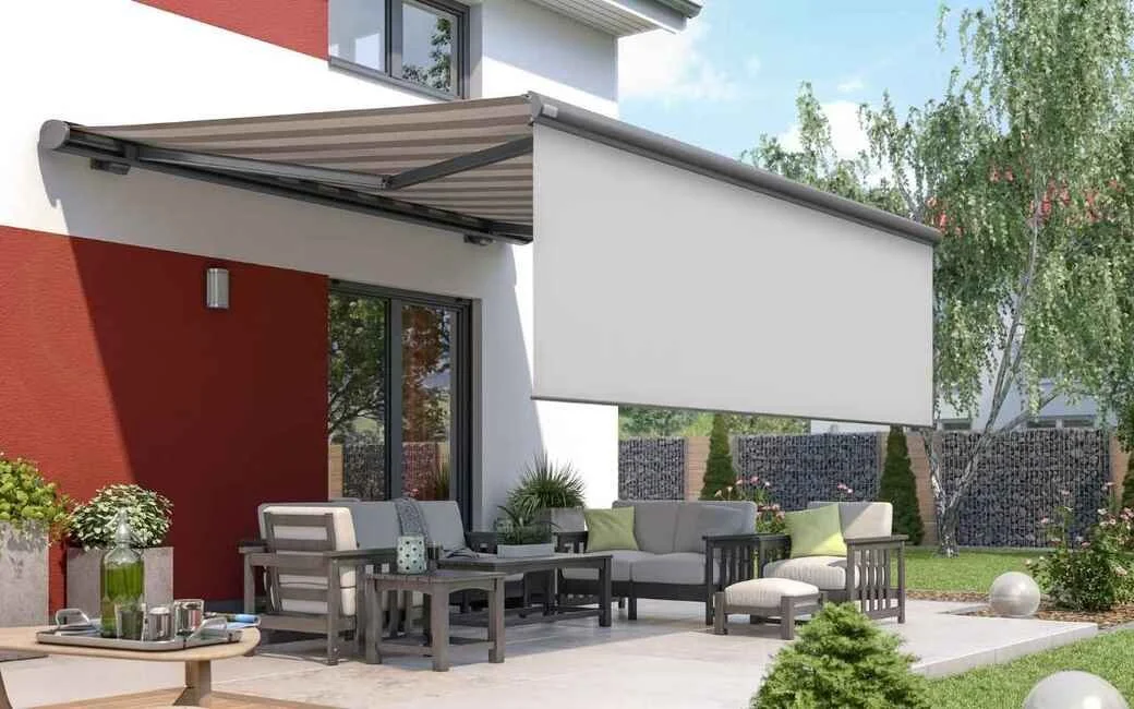 Gartenmöbel auf einer Terrasse mit einer Sonnenmarkise, im Hintergrund Bäume und ein Zaun.