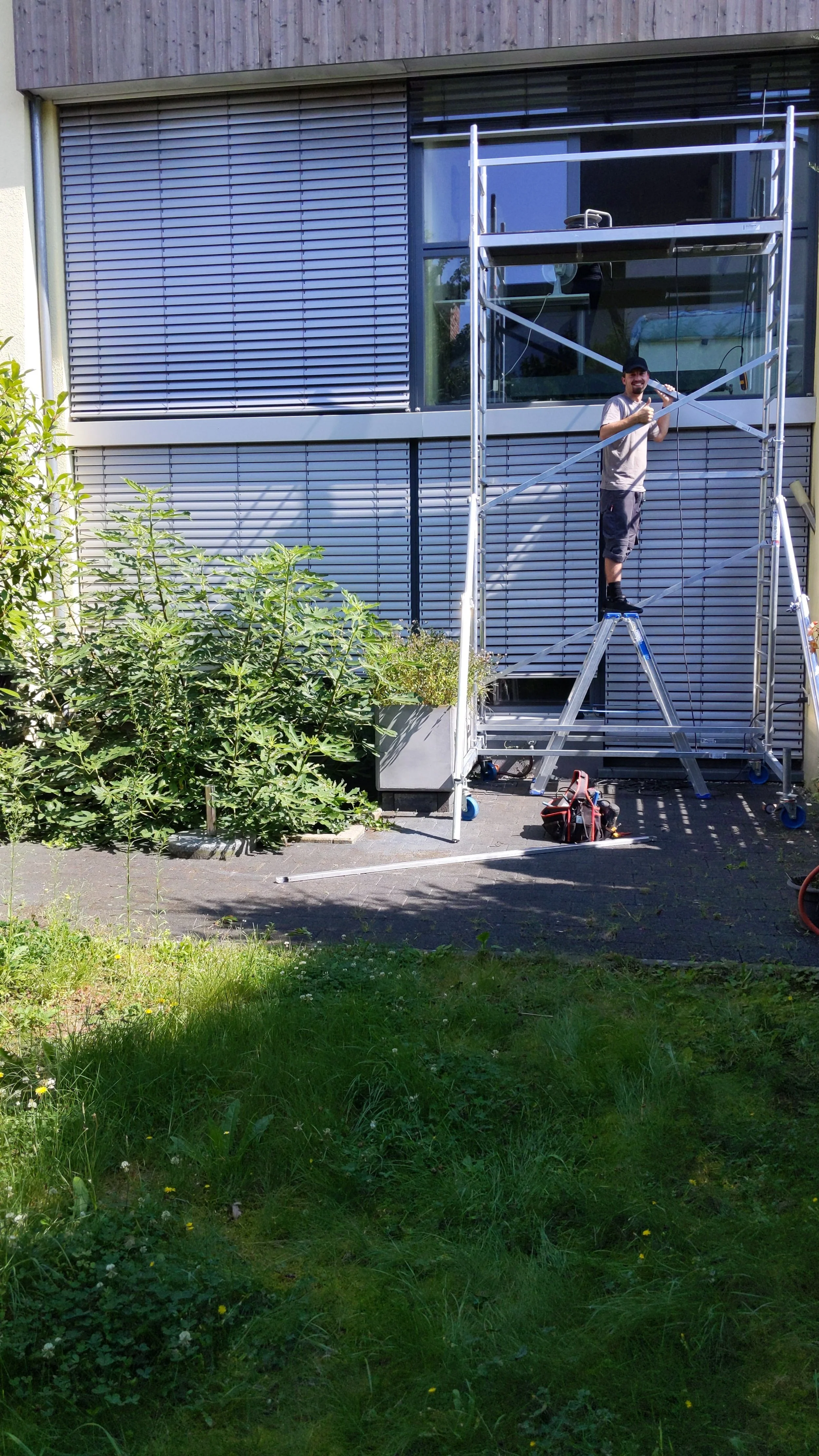 Ein Mann arbeitet auf einer Baustellengerüst vor einem modernen Gebäude mit verschatteten Fenstern.