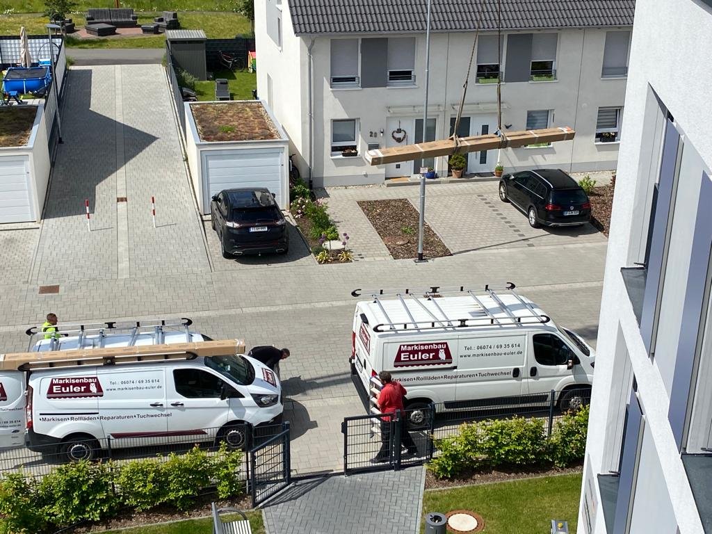 Zwei weißen Handwerksfahrzeuge der Firma Euler parken vor einer Wohnanlage. Zwei Arbeiter sind bei der Arbeit, ein Arbeiter trägt Holz auf die Leiter. Es ist ein Kran im Einsatz, um ein großes Holzstück zur Terrasse zu heben.