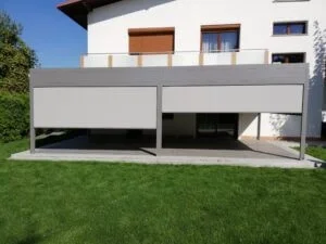 Ein modernes, zweistöckiges Haus mit einer offenen Terrasse auf der unteren Ebene, umgeben von grünem Rasen und einem Baum im Hintergrund.