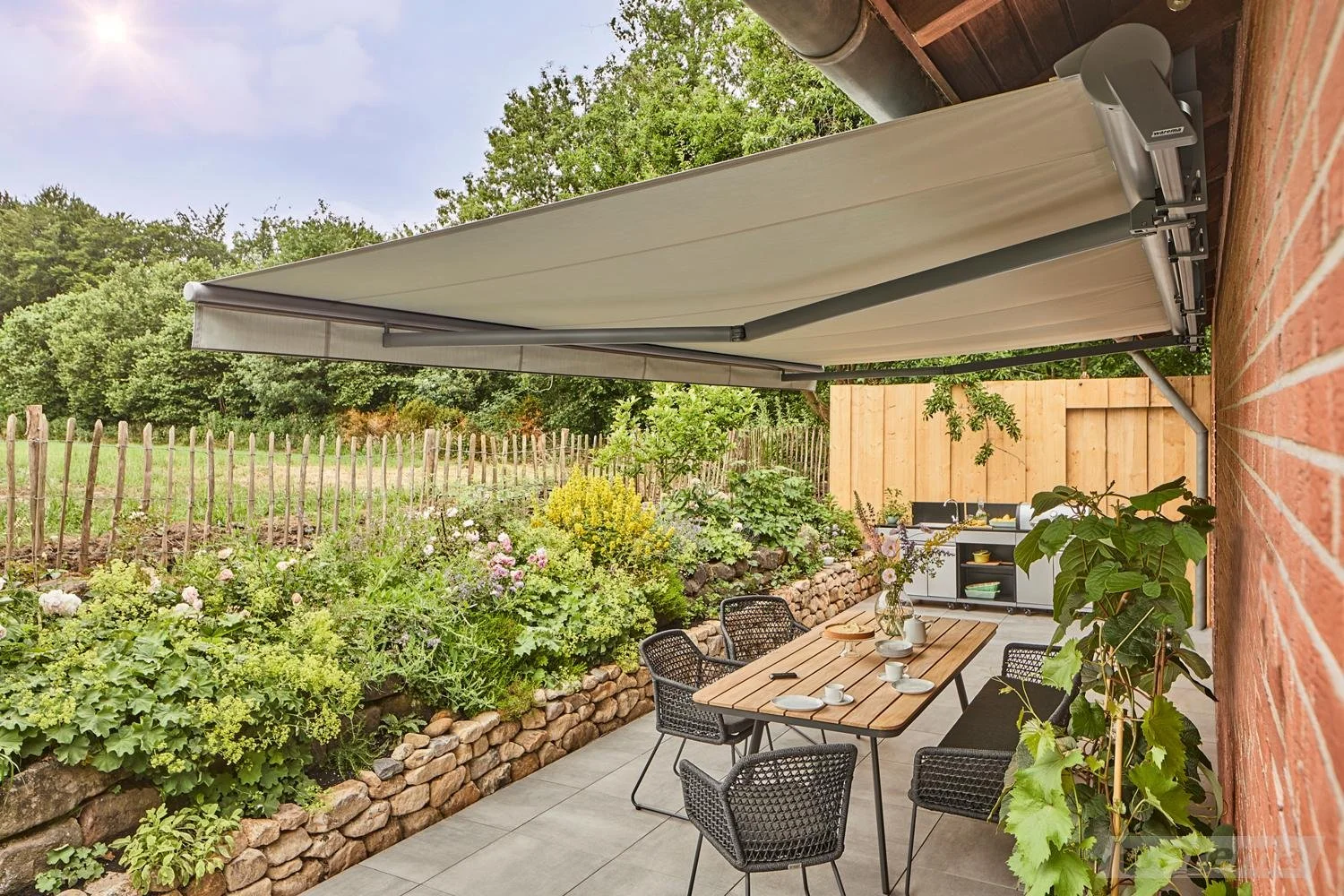 Ein gemütlicher Terrassenbereich mit einem rechteckigen Holztisch, sechs schwarzen Stühlen, gedeckt mit Geschirr, unter einer großen Markise. Es gibt bunte Blumen und Pflanzen in einem Steinbeet, einen kleinen Garten im Hintergrund und einen Holzzaun