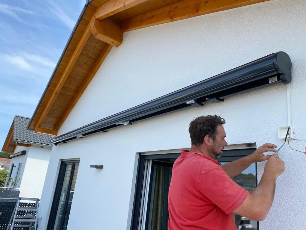 Ein Mann arbeitet an der Außenwand eines Hauses, wahrscheinlich installiert er eine elektrische Leitung. Das Haus hat weiße Wände, große Fenster und ein Satteldach mit Holzverkleidung.
