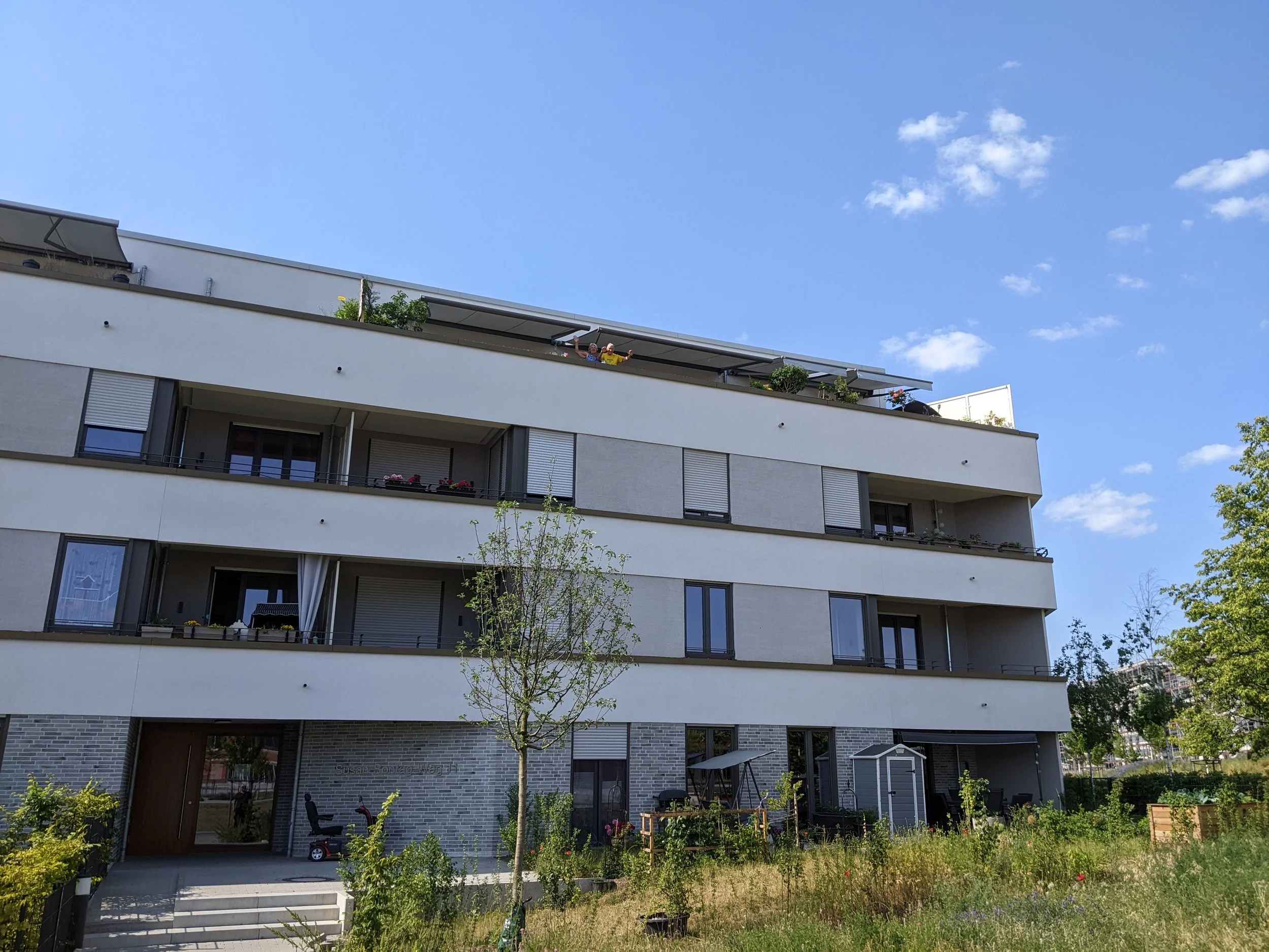 Mehrstöckiges modernes Wohngebäude mit Balkonen und Garten im Vordergrund, blaue Himmel mit wenigen Wolken.