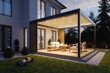 Modernes Haus mit einer verglasten Terrasse im Freien, abends beleuchtet, mit Garten und Bäumen im Hintergrund.