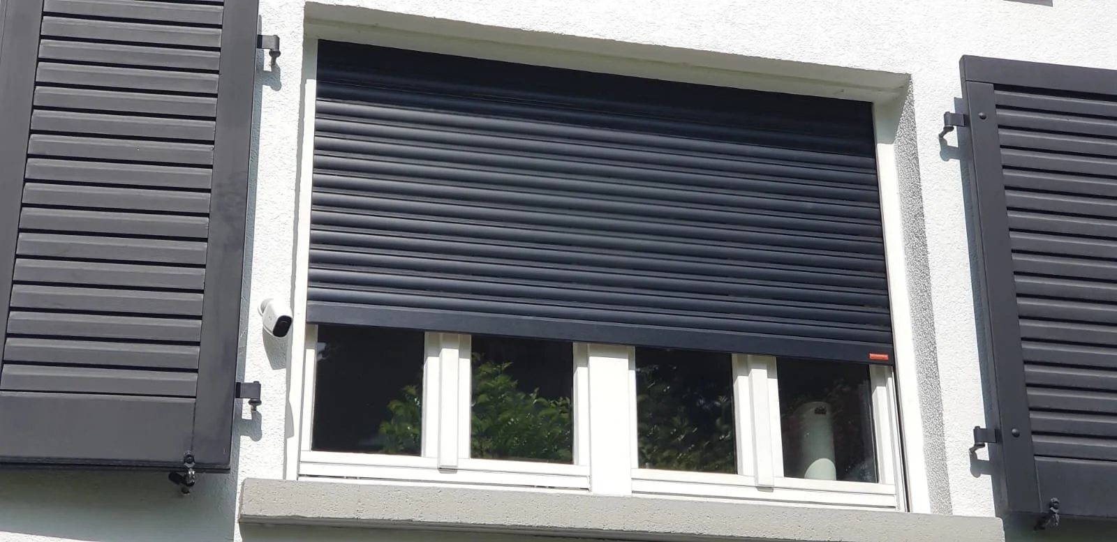 Fenster mit schwarzen Rollläden und geöffneten schwarzen Fensterläden an einer weißen Hauswand.