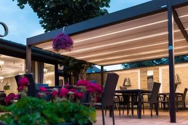 Ein modern eingerichteter Außenbereich eines Restaurants oder Cafés mit Tischen, Stühlen und eine Überdachung. Es sind Blumen und Pflanzen sichtbar, bei Tageslicht.