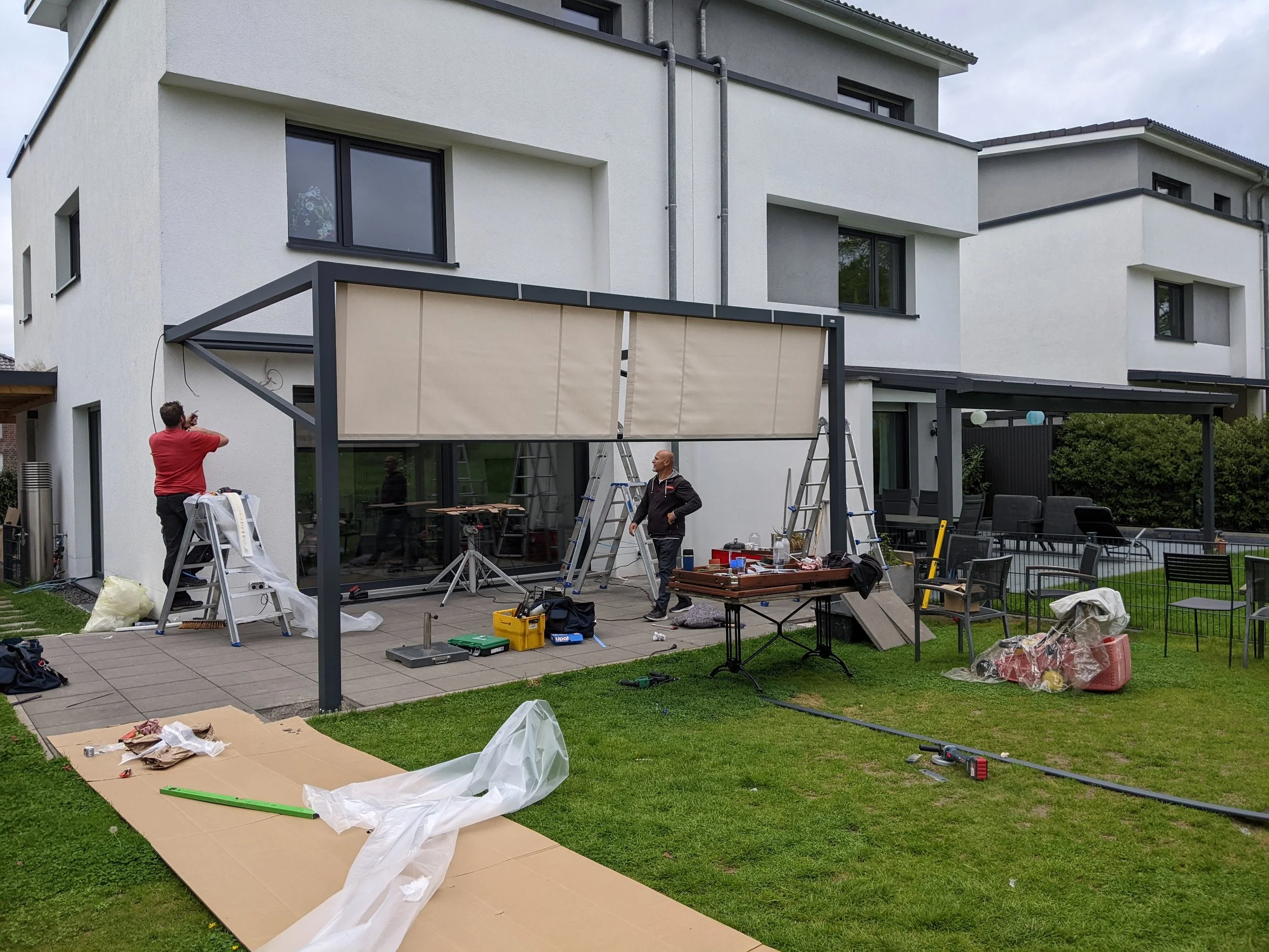 Arbeiten im Außenbereich eines modernen weißen Hauses. Zwei Männer installieren eine Sonnenschutzblende an einem Terrassendach, mit Werkzeugen und Baumaterialien auf Tischen und Bodengroßfolie. Es gibt einen Garten mit Gras, mehrere Stühle, und einen