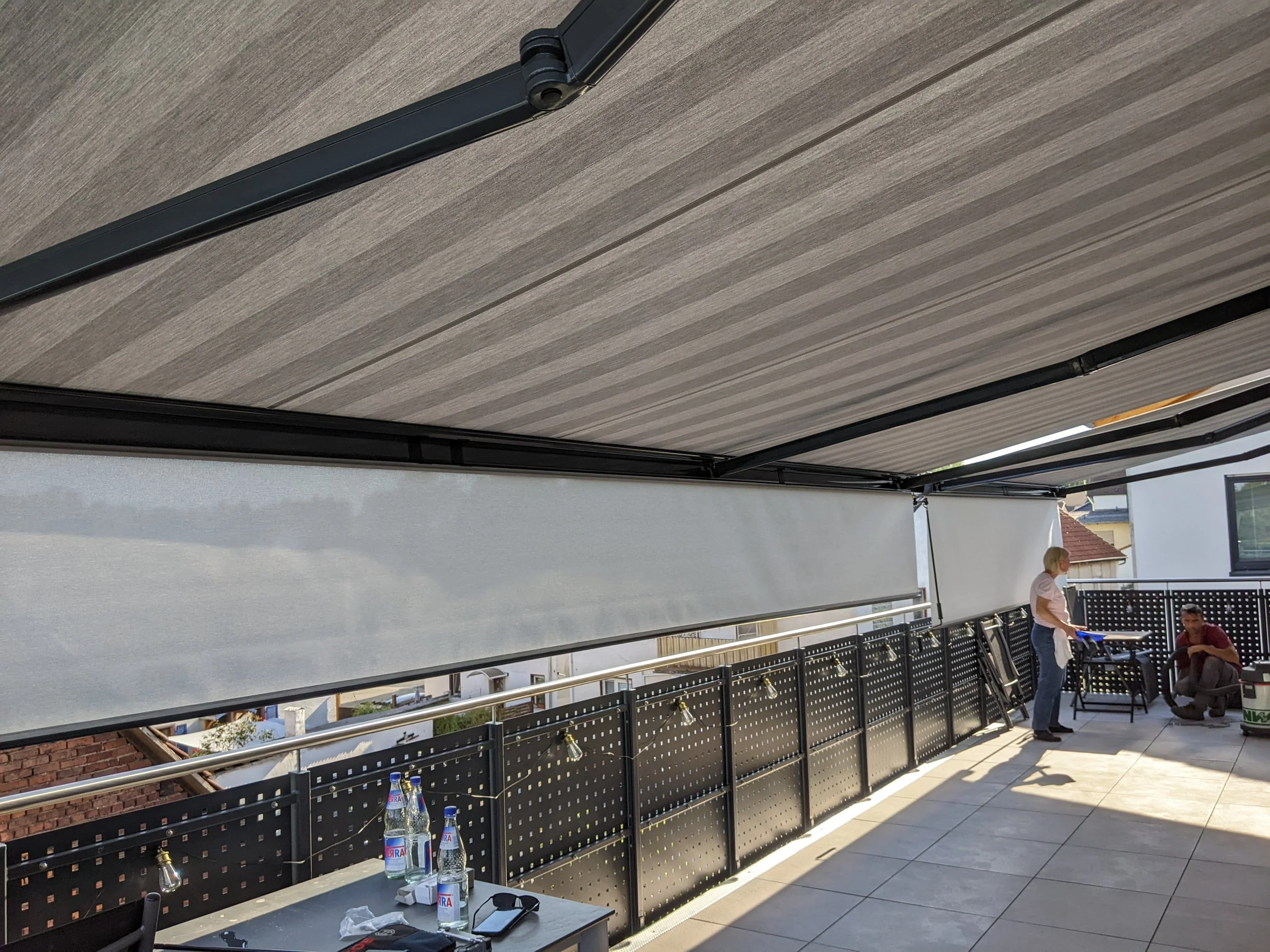 Blick auf eine ungenutzte Dachterrasse mit mehreren Personen, die an einem Grill arbeiten, und einem Tisch mit Getränken, mit Sonnenblenden und einem Stahlgeländer.