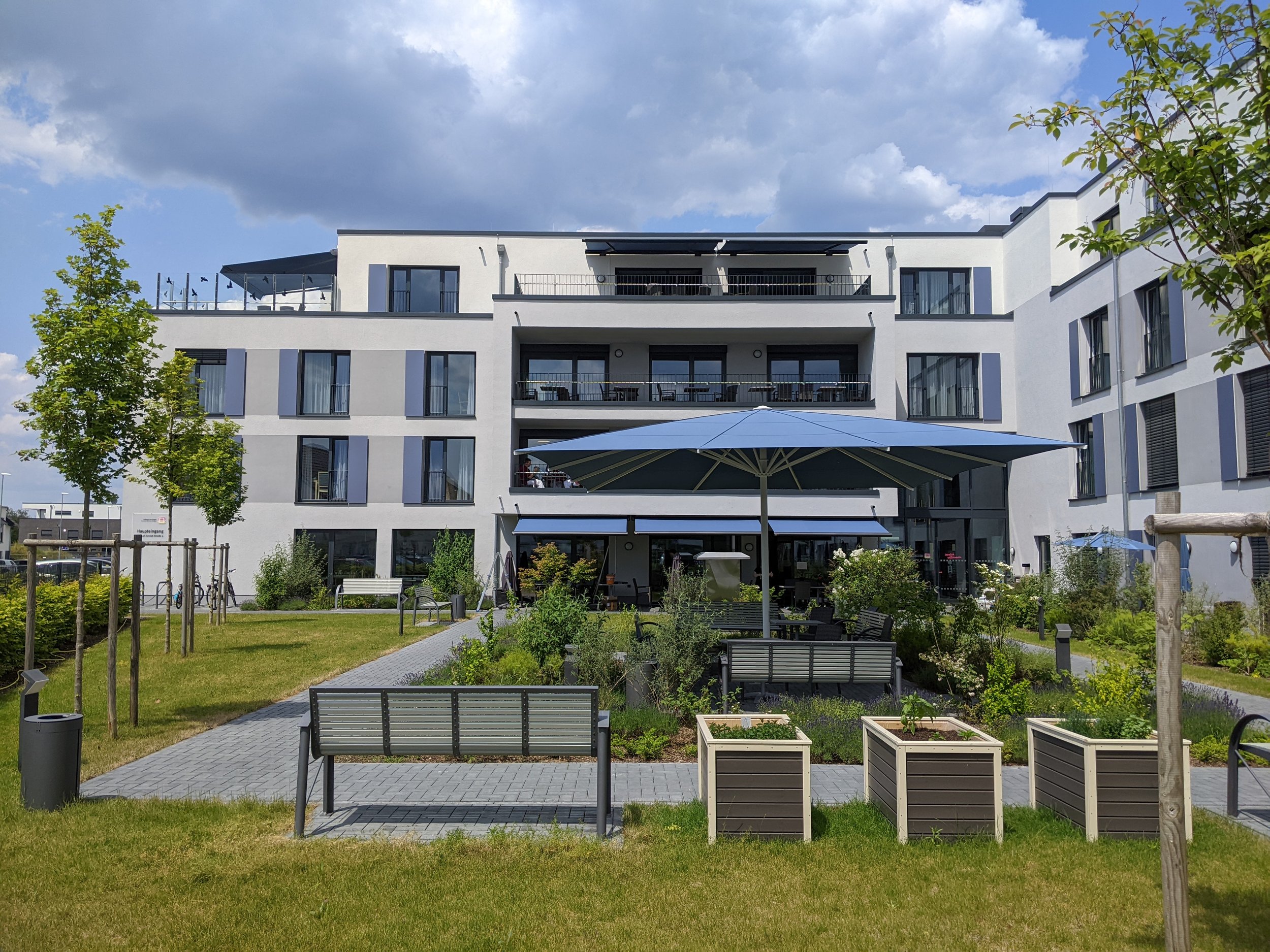 Moderner Mehrfamilienwohnblock mit Garten und Terrasse, Sonnenschirm, Bänke und bepflanzte Hochbeete vor blauen Himmel mit Wolken.