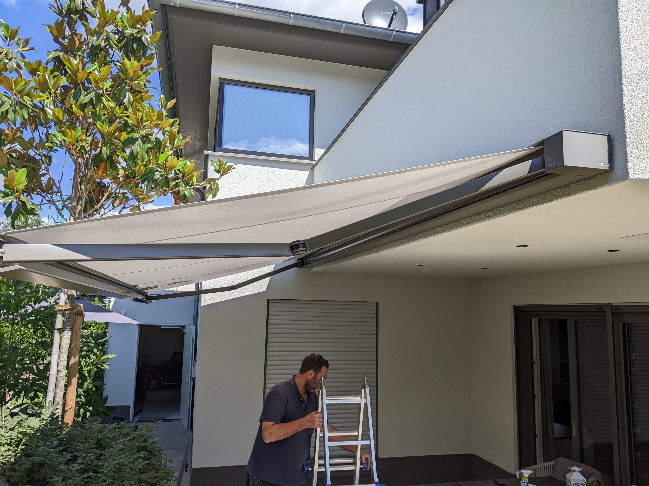 Ein Mann steht auf einer Leiter auf einer Terrasse und arbeitet an einem elektrischen Sonnenschutzschirm, der an der Außenwand eines Hauses angebracht ist.