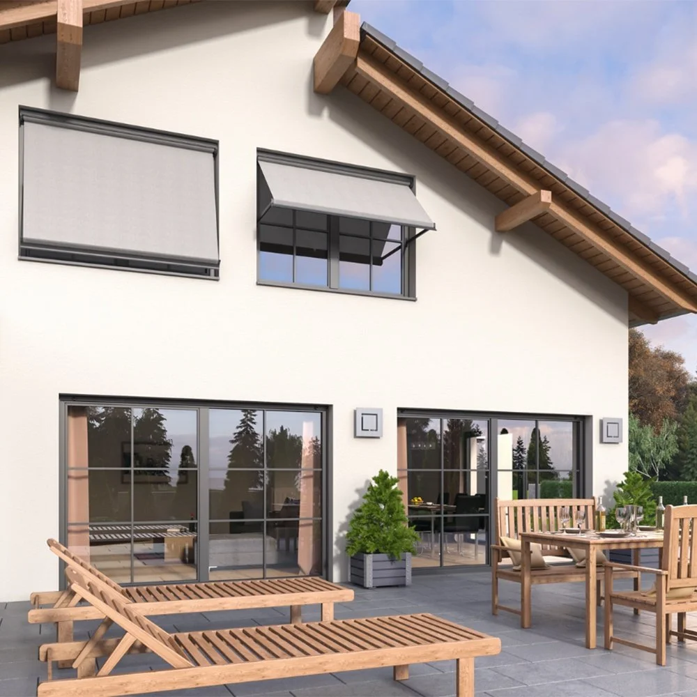 Ein modernes Haus mit großen Fenstern und einem Balkon. Im Vordergrund befinden sich Gartenmöbel, darunter ein Tisch, Stühle und Liegen, auf einer Terrasse mit grauen Fliesen. Es gibt auch grüne Pflanzen auf der Terrasse.
