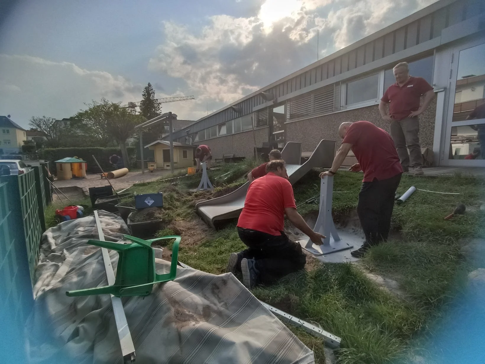 Mitarbeiter installieren eine Rutsche im Garten eines Gebäudes. Es sind mehrere Personen bei der Arbeit, mit Werkzeugen und Materialien, um die Rutsche aufzustellen.