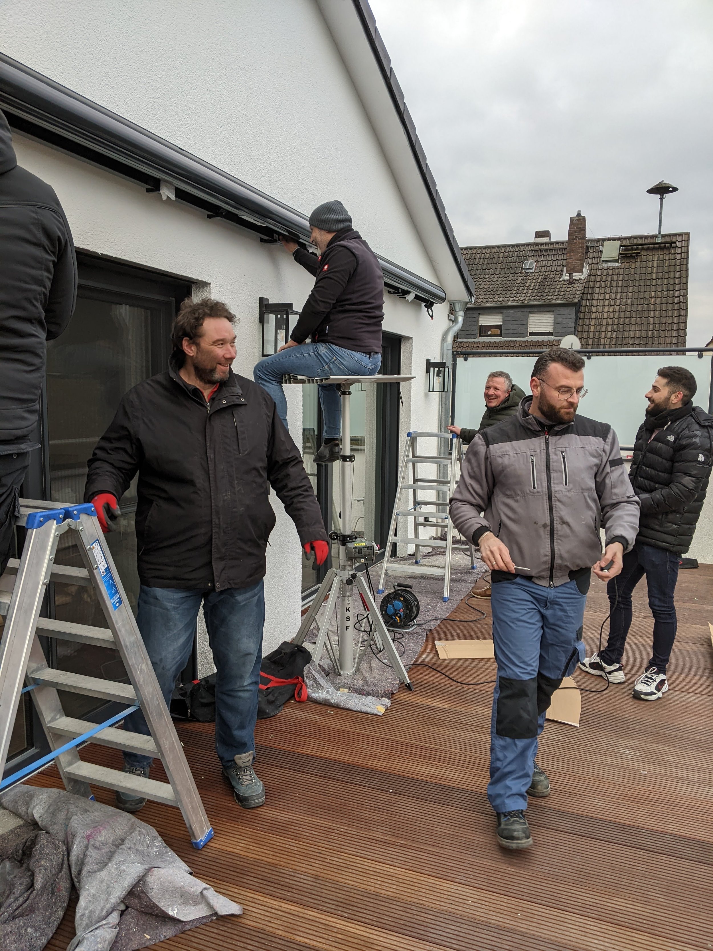 Gruppe von Menschen arbeitet auf einer Dachterrasse mit Werkzeug und Leiter, während sie wahrscheinlich eine Renovierungs- oder Bauarbeit durchführen.
