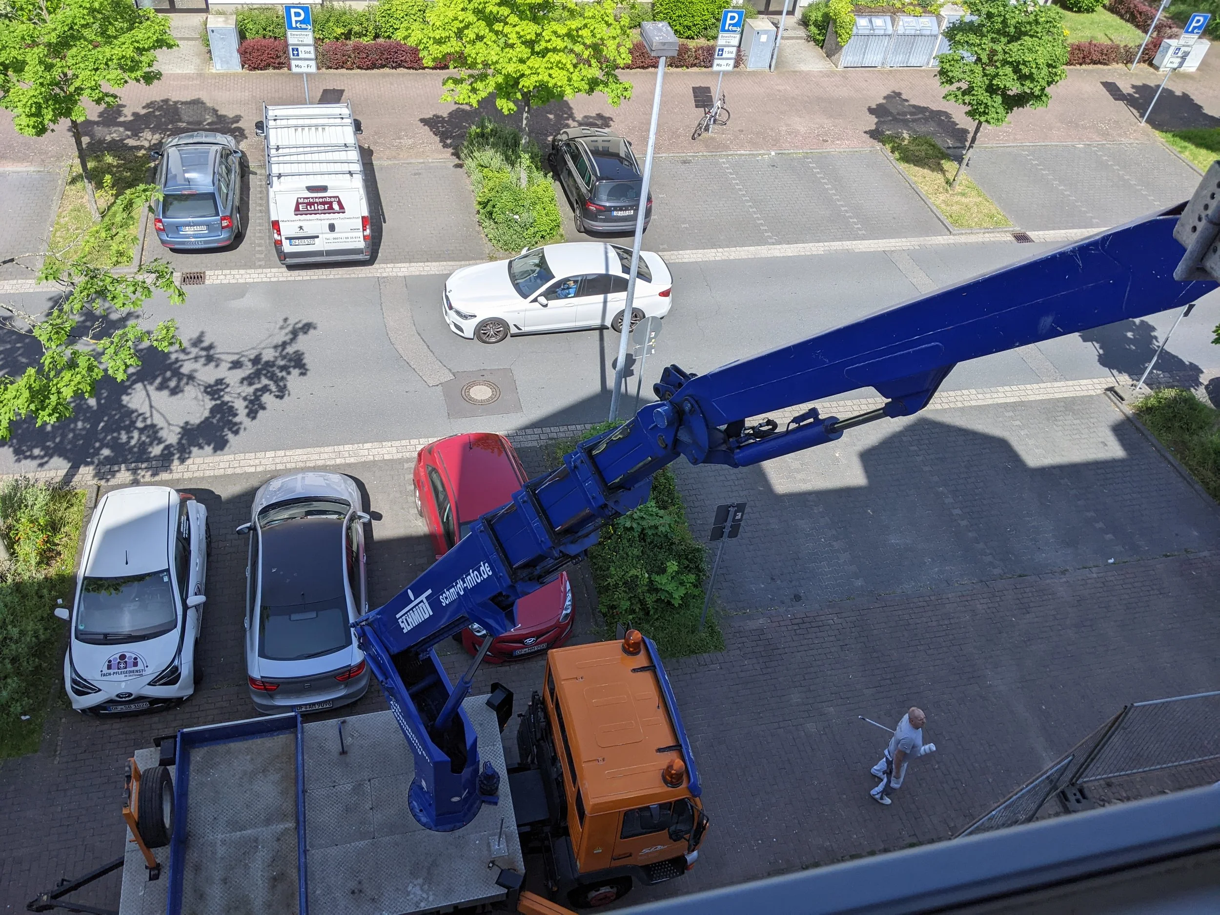 Blick von oben auf eine Straße mit parkenden Autos und einem aufgestellten Kran mit orangefarbenem Fahrgestell. Es sind Bäume, Parkplätze und eine Person zu sehen, die einen weißen Lappen in der Hand hält.