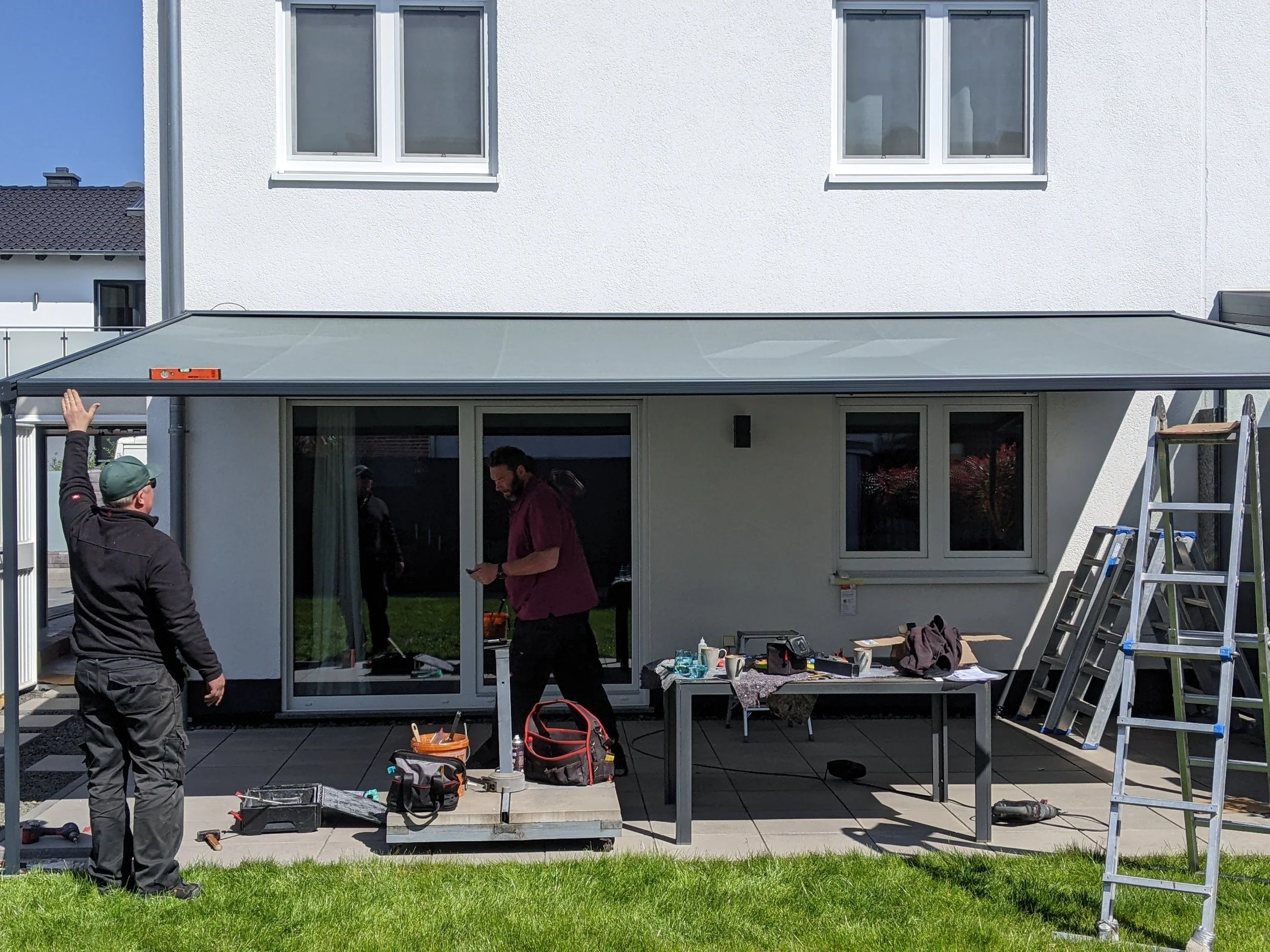 Zwei Männer bauen einen Überdachung im Garten eines Hauses. Es sind Werkzeuge und Baumaterialien auf dem Tisch and im Boden.