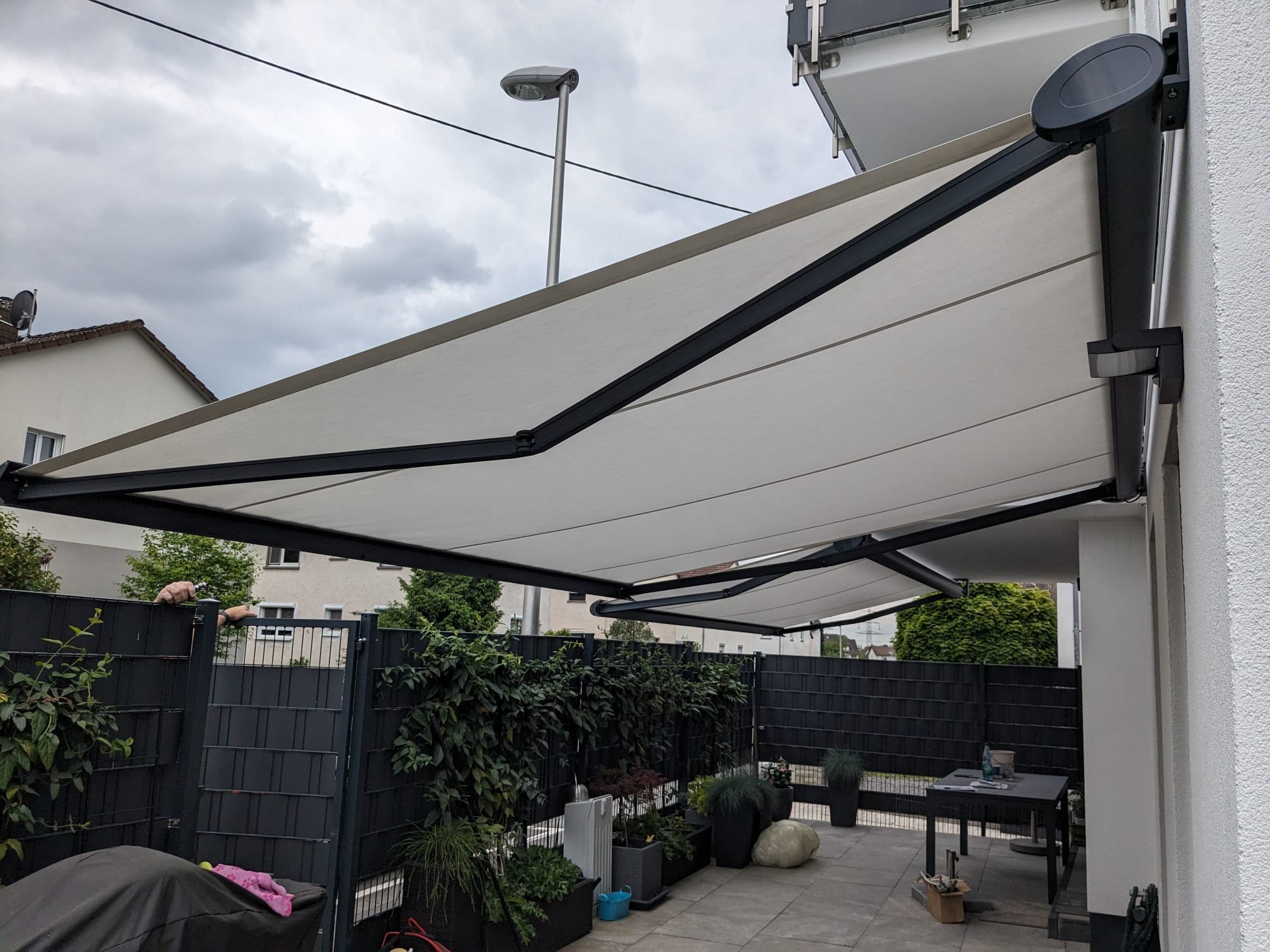 Automatischer Sonnenschutz im Garten, halb aufgefahren, auf einem Balkon vor einer weißen Hauswand.