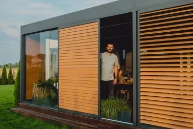 Mann steht in einem modernen Haus mit großen Glastüren und Holzlamellen, blickt nach draußen.