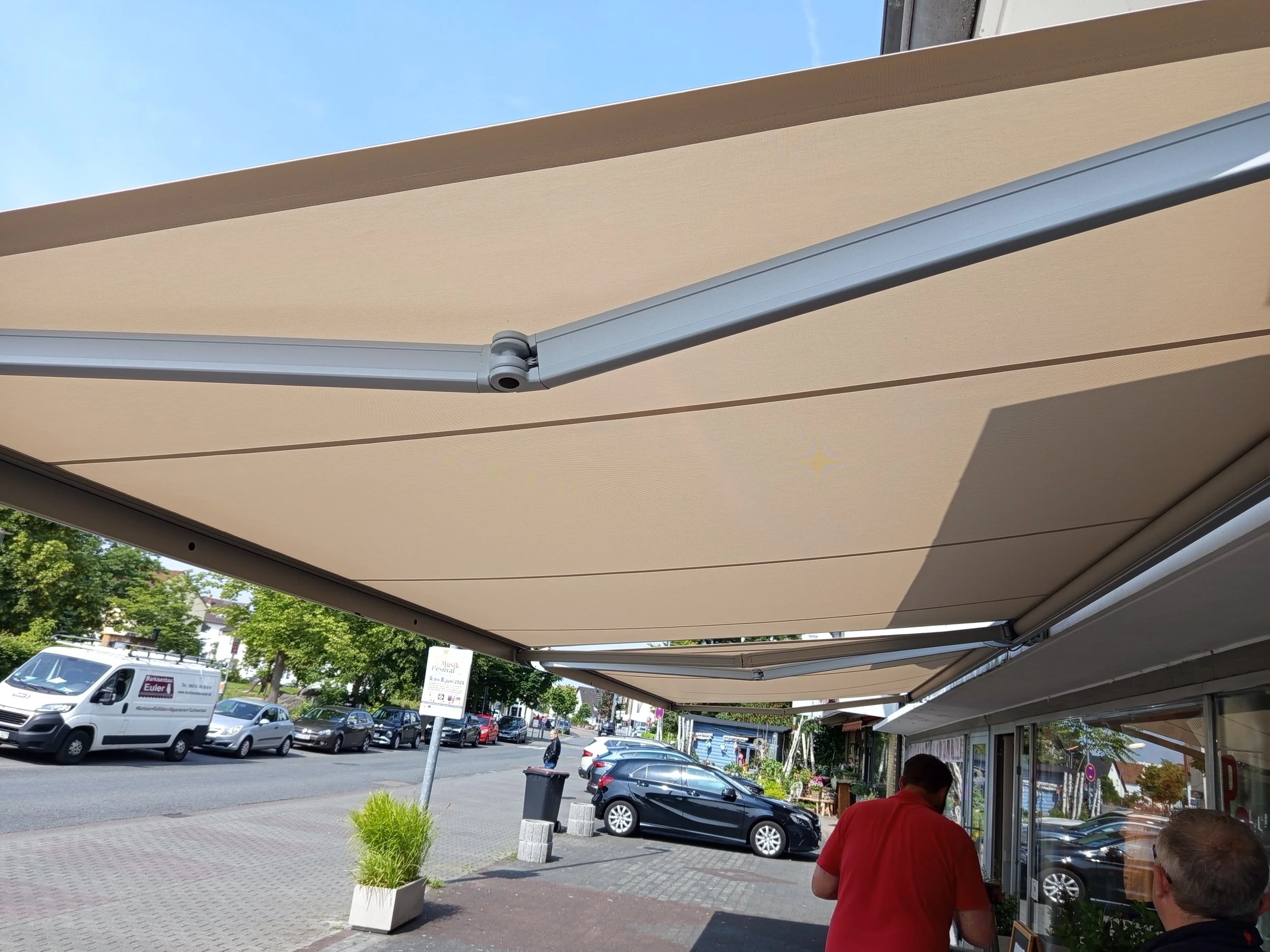 Sonnenschutzmarkise über einem Außencafé mit Blick auf die Straße, mehrere Autos und Menschen, die draußen sitzen.