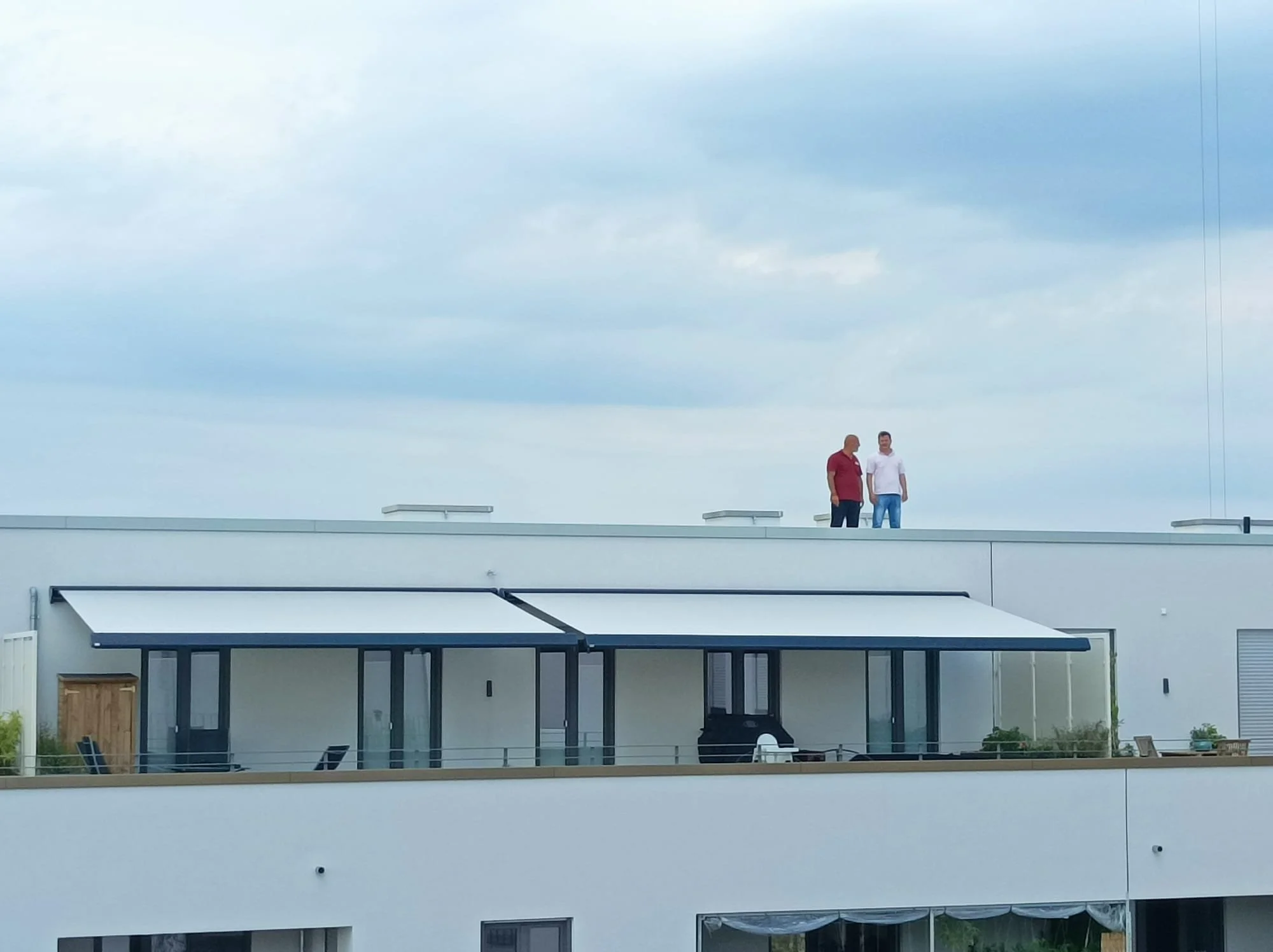 Zwei Männer stehen auf dem Dach eines modernen Gebäudes unter einem bewölkten Himmel.