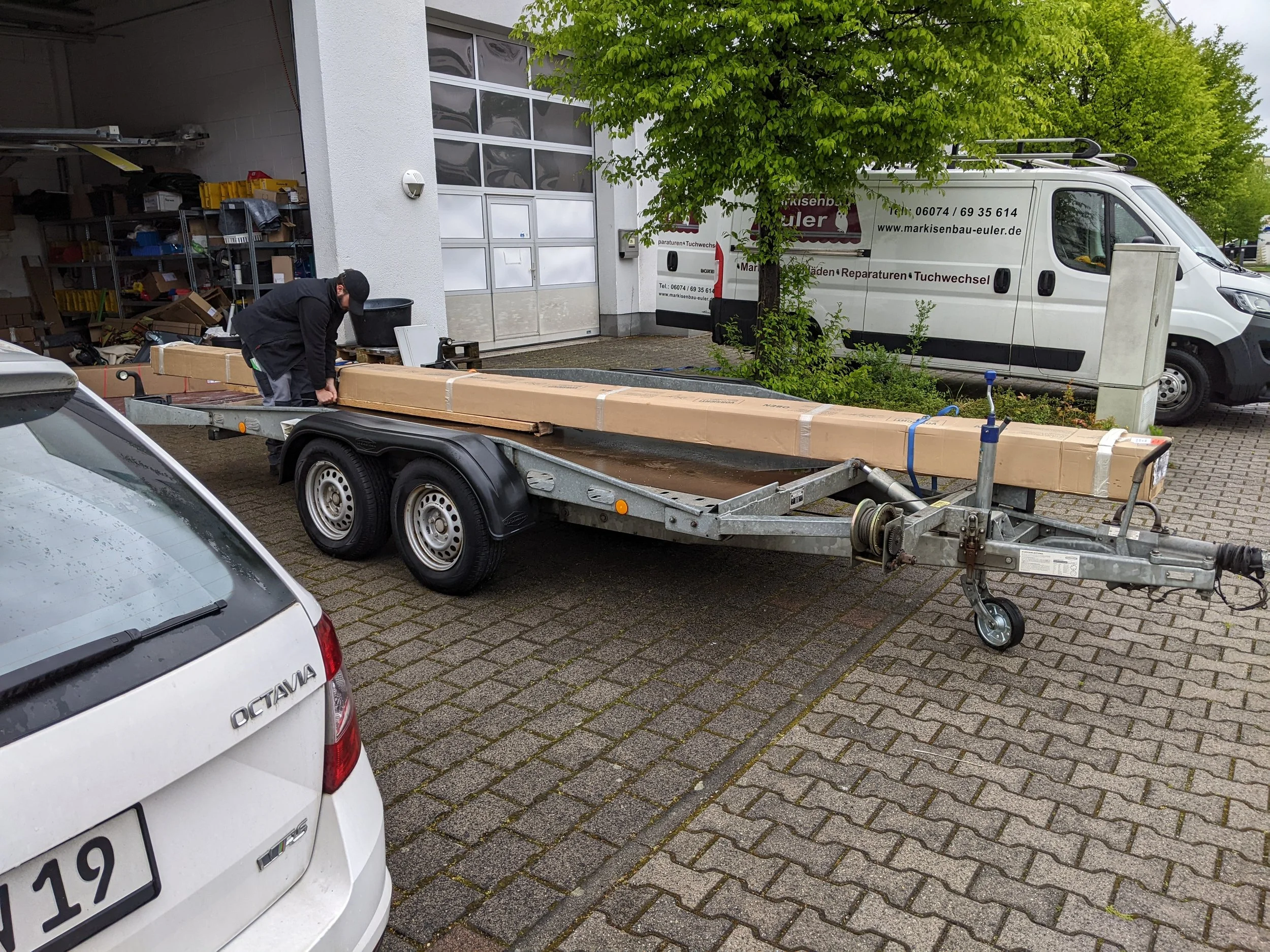 Ein Mann läuft auf einem flachen Anhänger, der ein langes Paket transportiert, vor einer Werkstatt. Im Hintergrund stehen Fahrzeuge und die Werkstatttür ist geöffnet, es gibt Werkzeuge und Regale im Inneren.
