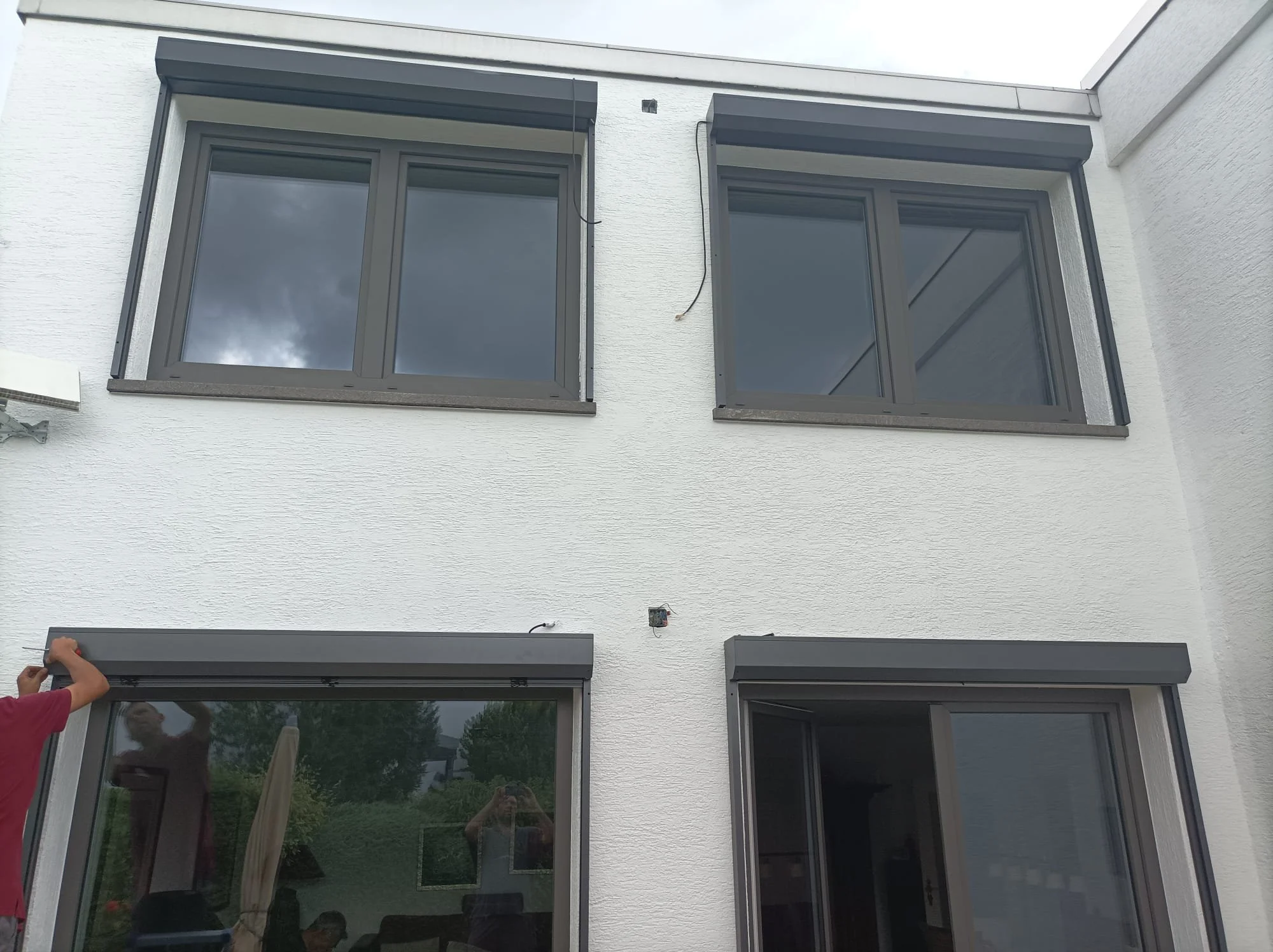 Auf einem weißen Haus sind vier große Fenster, zwei im oberen Stock und zwei im unteren. Es scheint, als ob jemand die Rollläden eines Fensters im unteren Stock präpariert oder installiert.