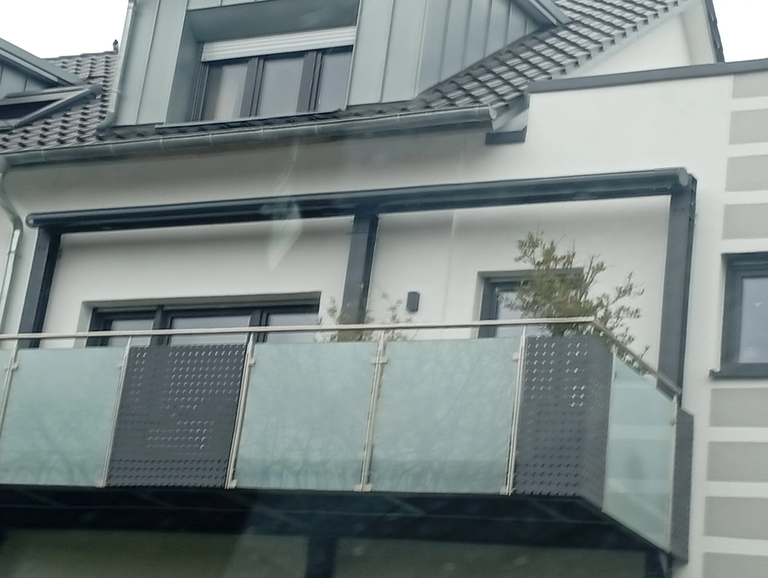Mehrstöckiges modernes Wohngebäude mit Balkon, Dachfenster und einer kleinen Pflanze auf dem Balkon, im Hintergrund ein Teil des Daches und anderes Gebäude. 