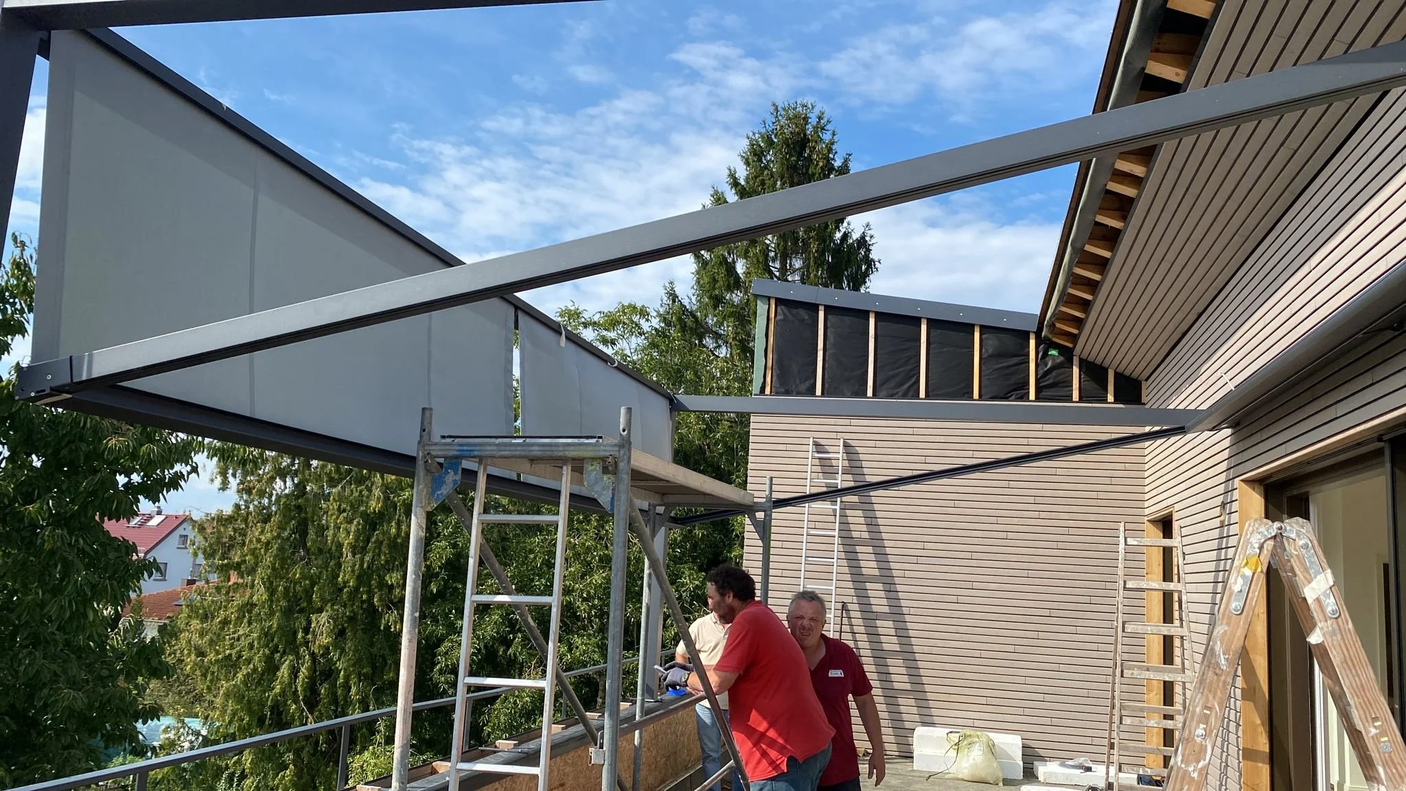 Bauarbeiter errichten eine Dachterrasse an einem modernen Haus, arbeiten mit Gerüsten und Leitern bei gutem Wetter.