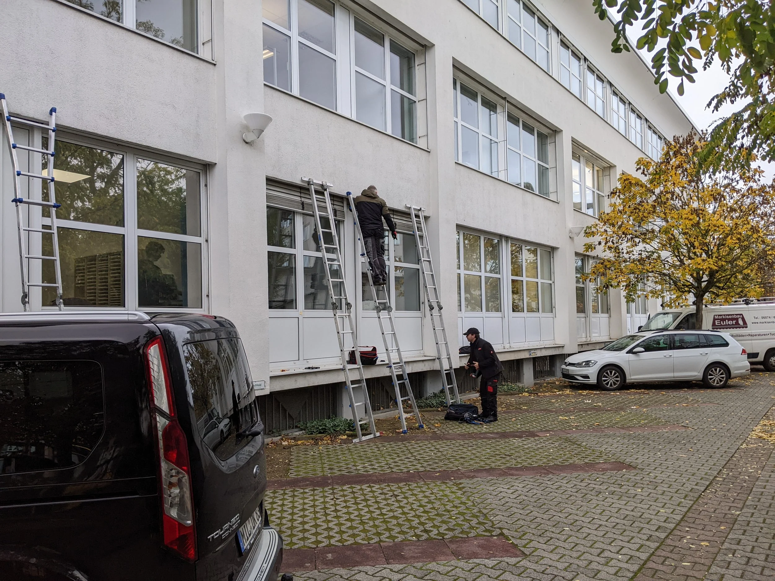 Zwei Männer stehen auf dem Bürgersteig vor einem weißen Gebäude mit großen Fenstern. Sie bereiten sich auf die Arbeit vor, mit Leitern vor dem Gebäude. Es sind einige parkende Autos und ein Baum mit herbstlichen Blättern sichtbar.