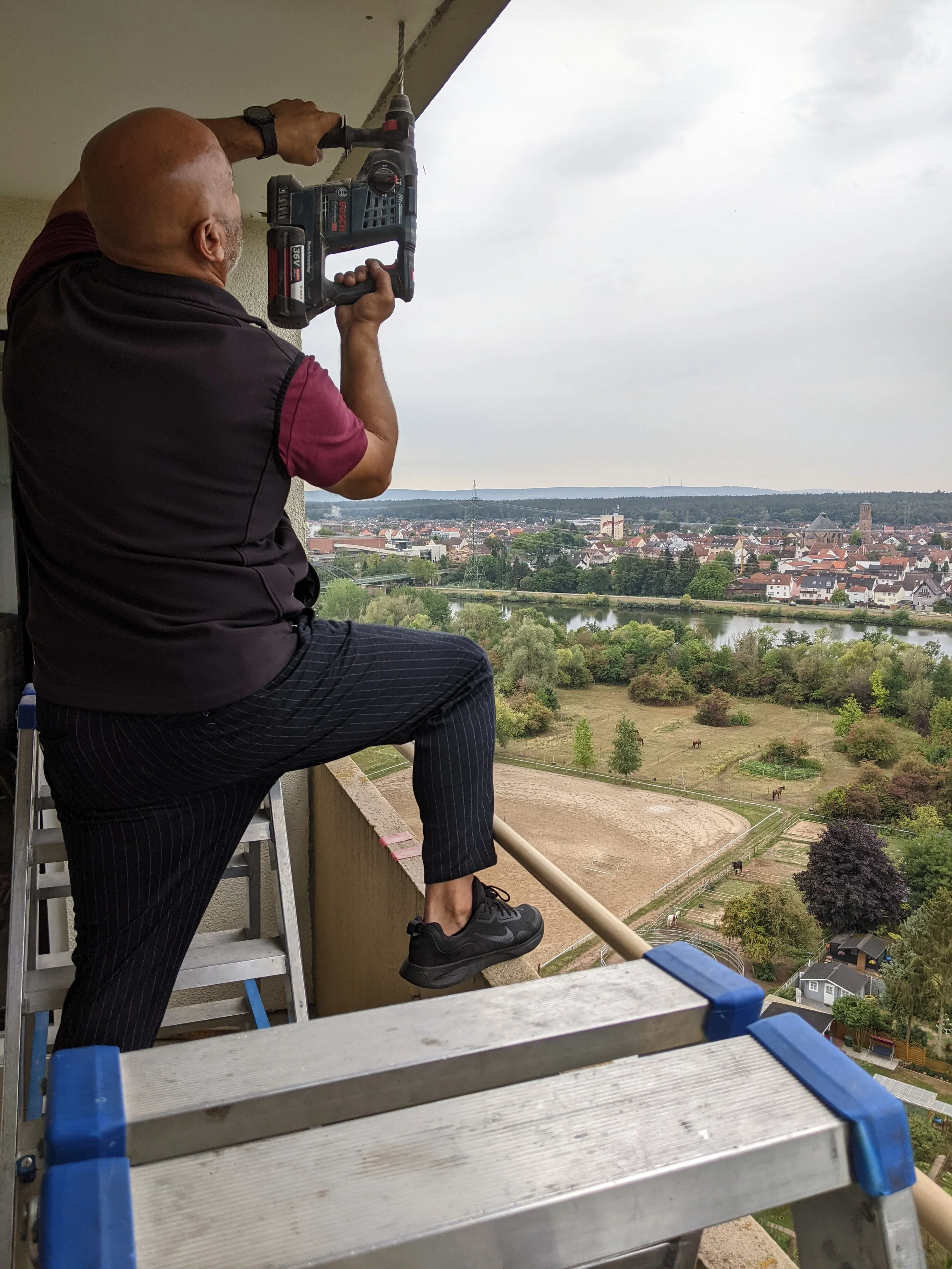Ein Mann steht auf einer Leiter auf einem Balkon und benutzt eine Bohrmaschine, um an die Decke zu bohren. Im Hintergrund ist eine Stadt mit Fluss und grünen Flächen zu sehen.