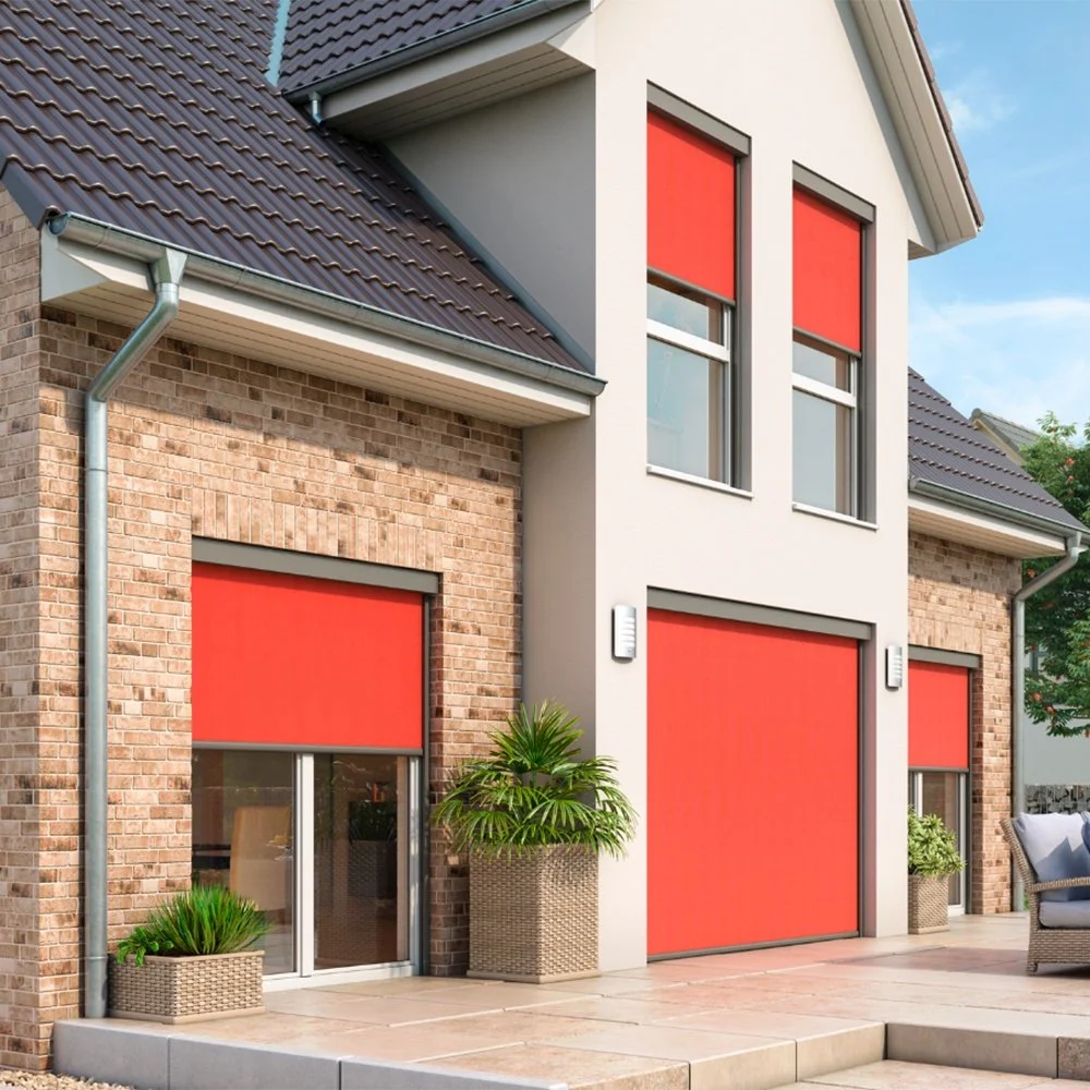 Modernes Haus mit rot-blauen Markisen an den Fenstern und einem großen roten Rolltor, braune Ziegelwände, weiße Fassadenteile, grünen Pflanzen in Körben, grauem Sofa im Außenbereich und blauen Himmel.